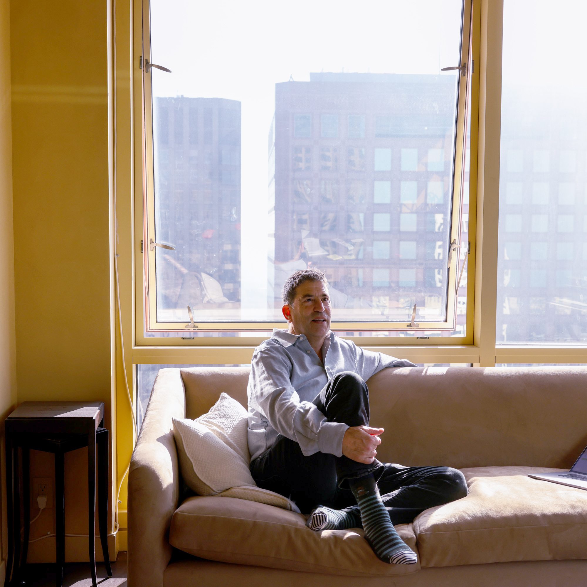 A man in a light blue shirt and striped socks sits on a beige couch, looking relaxed. Sunlight streams through large windows, with city buildings visible outside.