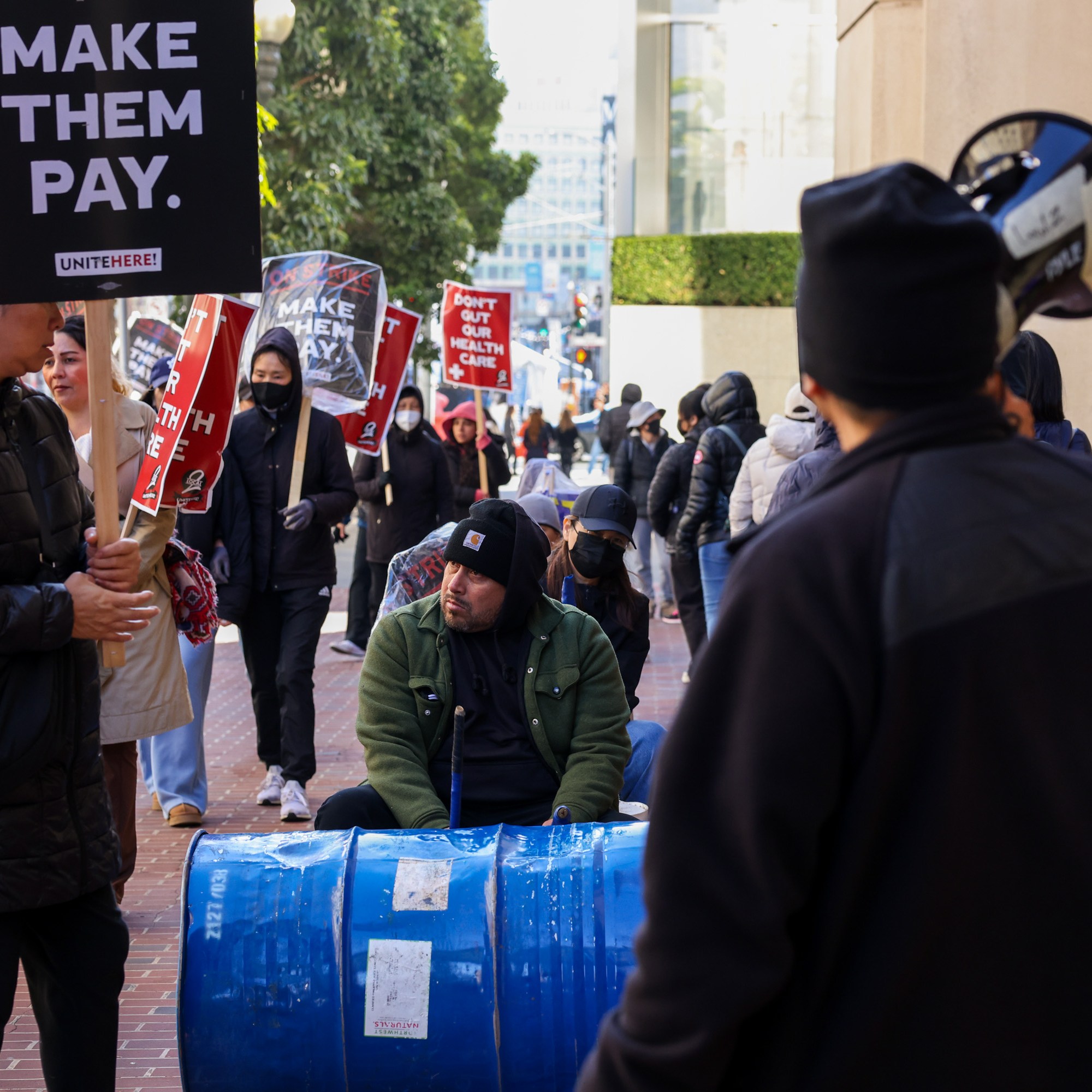 A group of people protesting, holding signs that say "DON'T GUT OUR HEALTH CARE" and "MAKE THEM PAY." One person sits near a blue barrel.