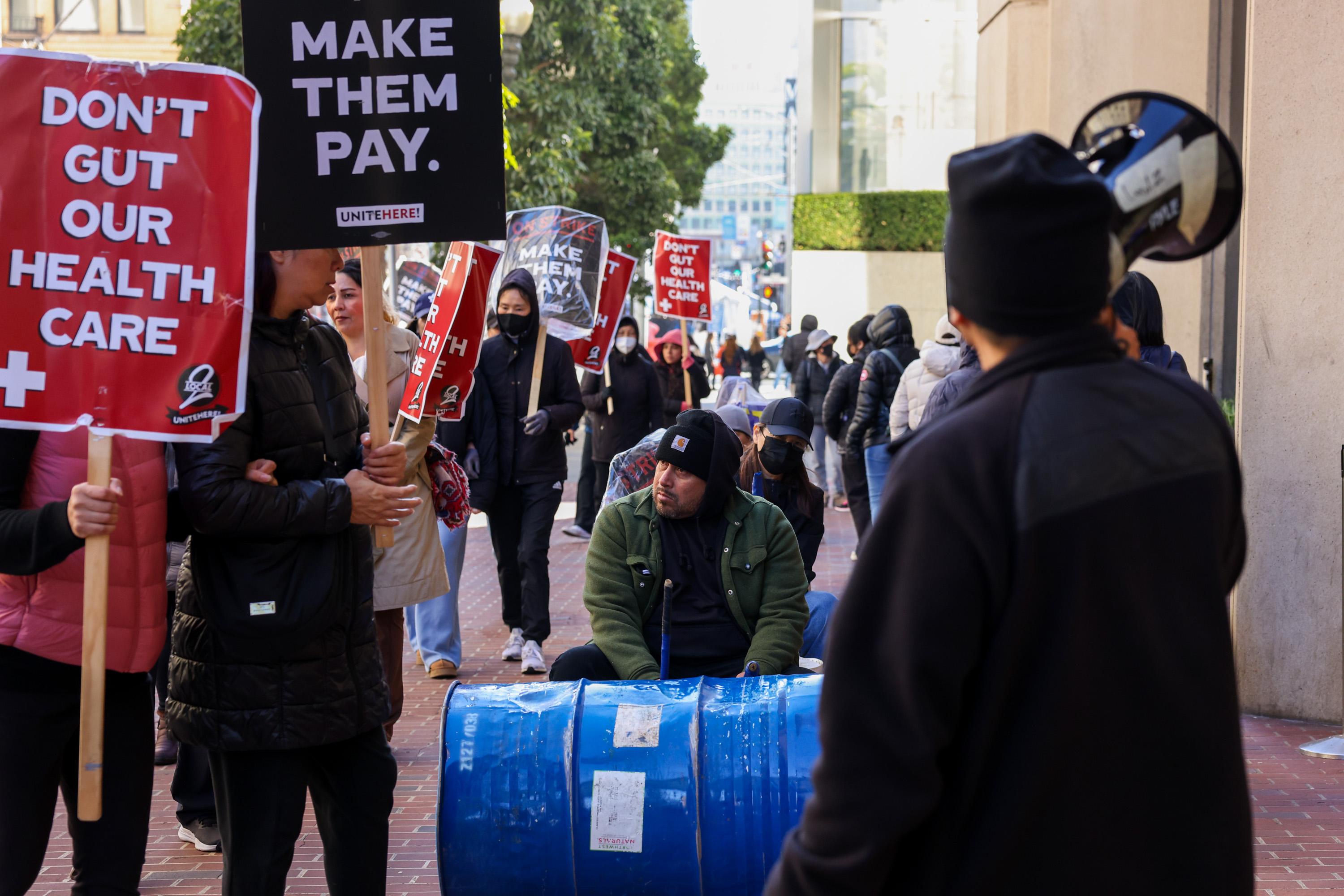 A group of people protesting, holding signs that say "DON'T GUT OUR HEALTH CARE" and "MAKE THEM PAY." One person sits near a blue barrel.