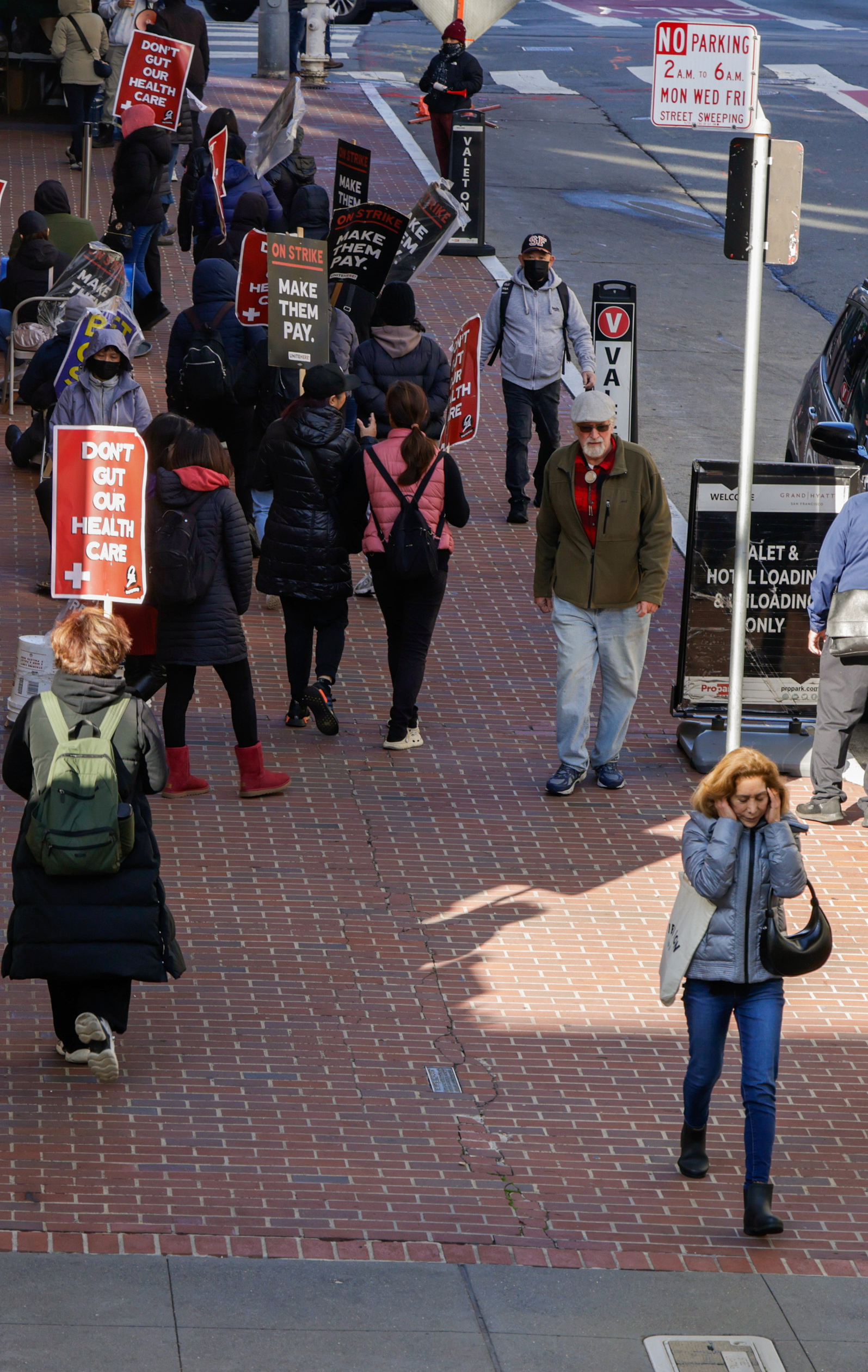 A group of people is marching on a brick sidewalk, holding signs that say &quot;Make Them Pay&quot; and &quot;Don’t Cut Our Health Care&quot; during a protest.