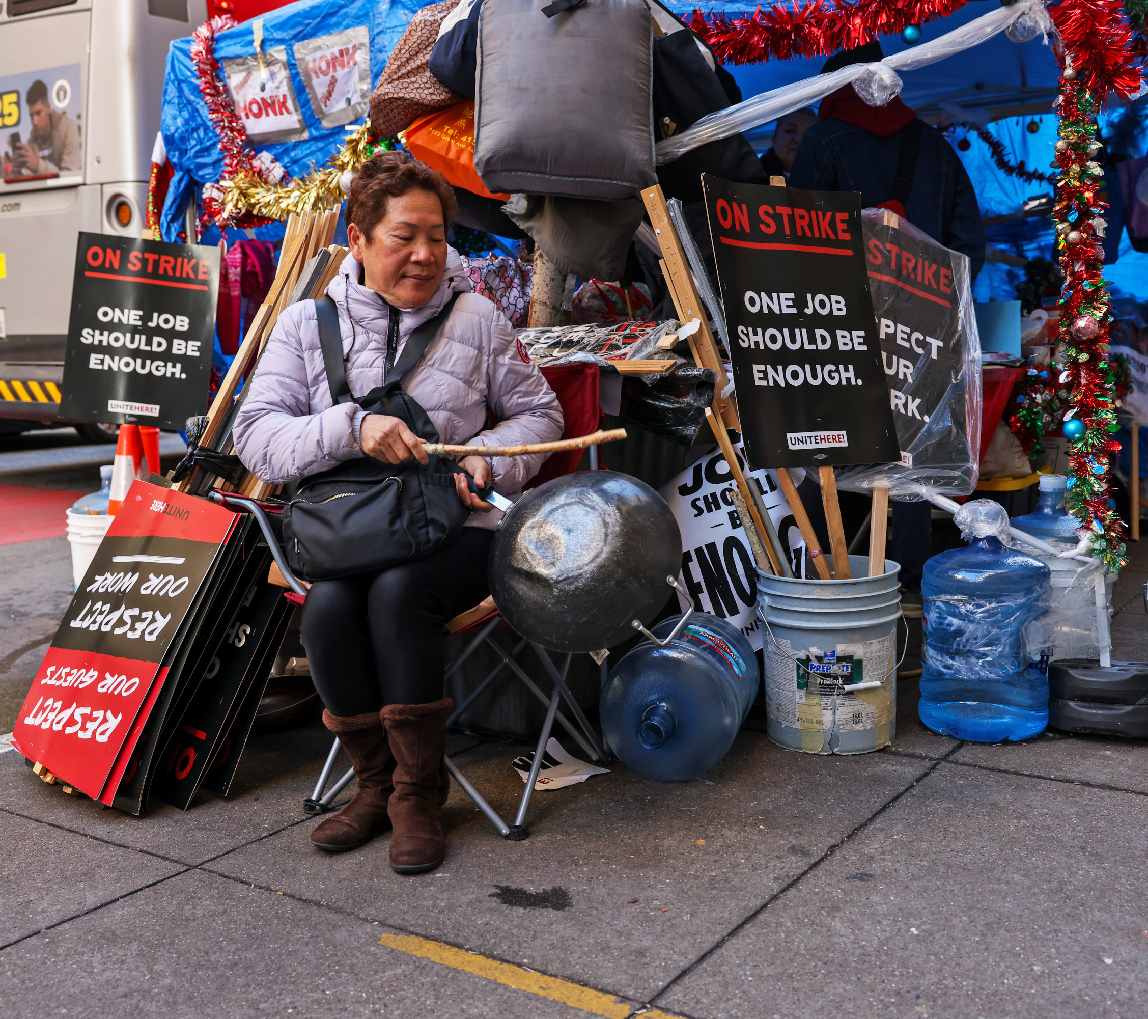 A person sits at a strike protest site with signs that read &quot;One job should be enough.&quot; They hold a drumstick and a large pot amidst colorful decorations.