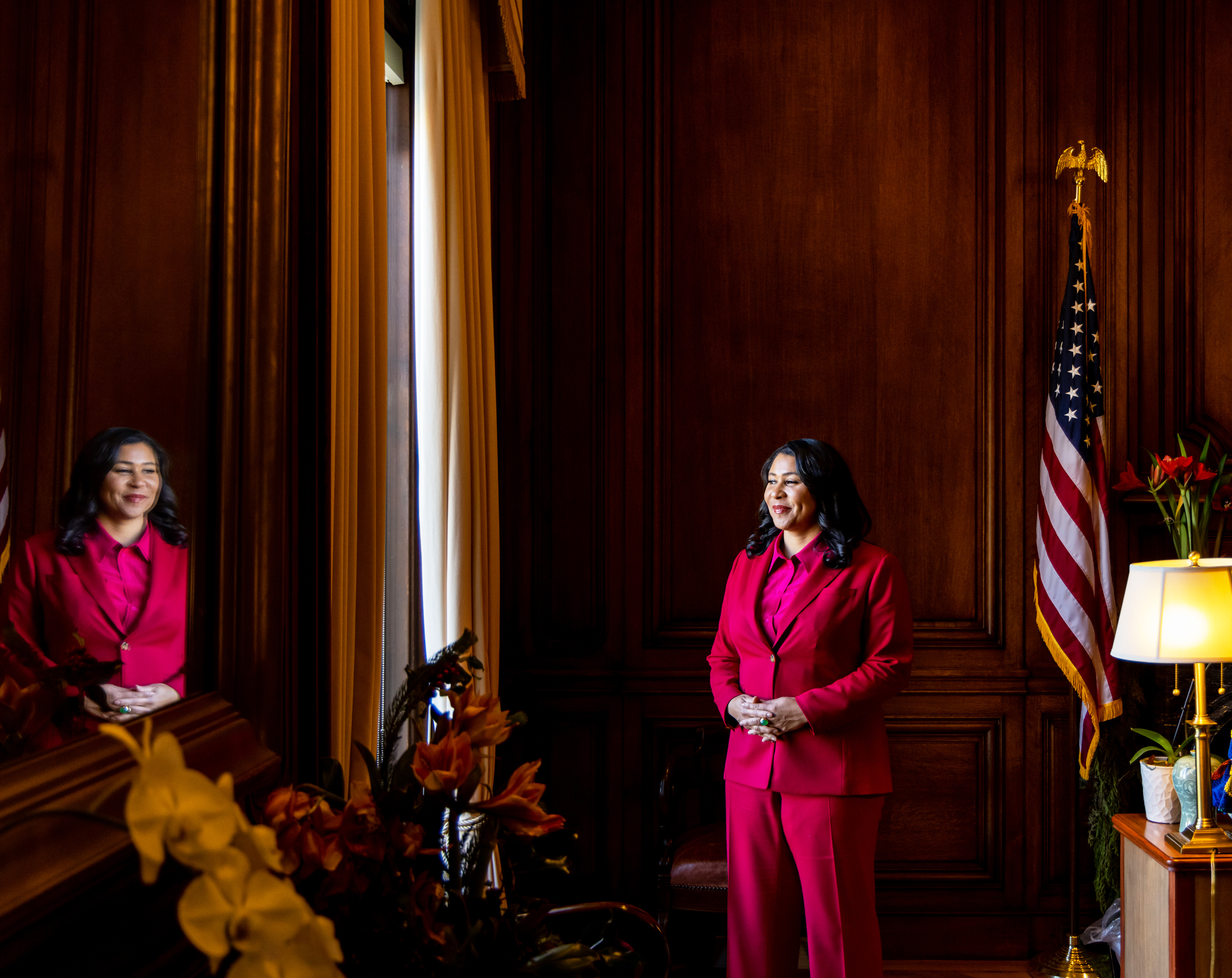 A woman in a bright pink suit stands confidently in a wood-paneled room. She is reflected in a mirror. An American flag and flowers are nearby.