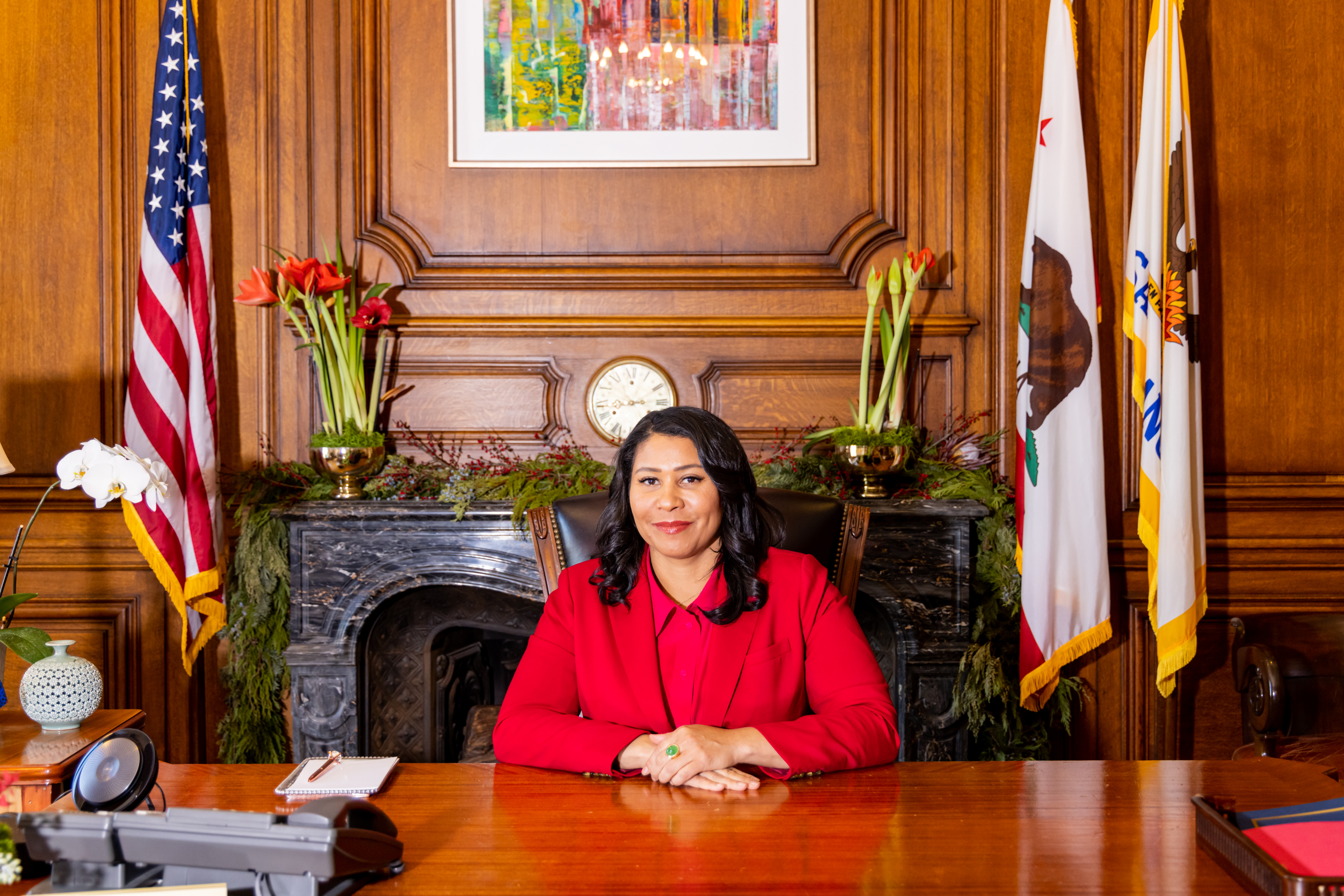 A person in a red outfit sits at a wooden desk in an ornate room. Behind them are flags, a clock, and flowers, with a colorful painting on the wall.