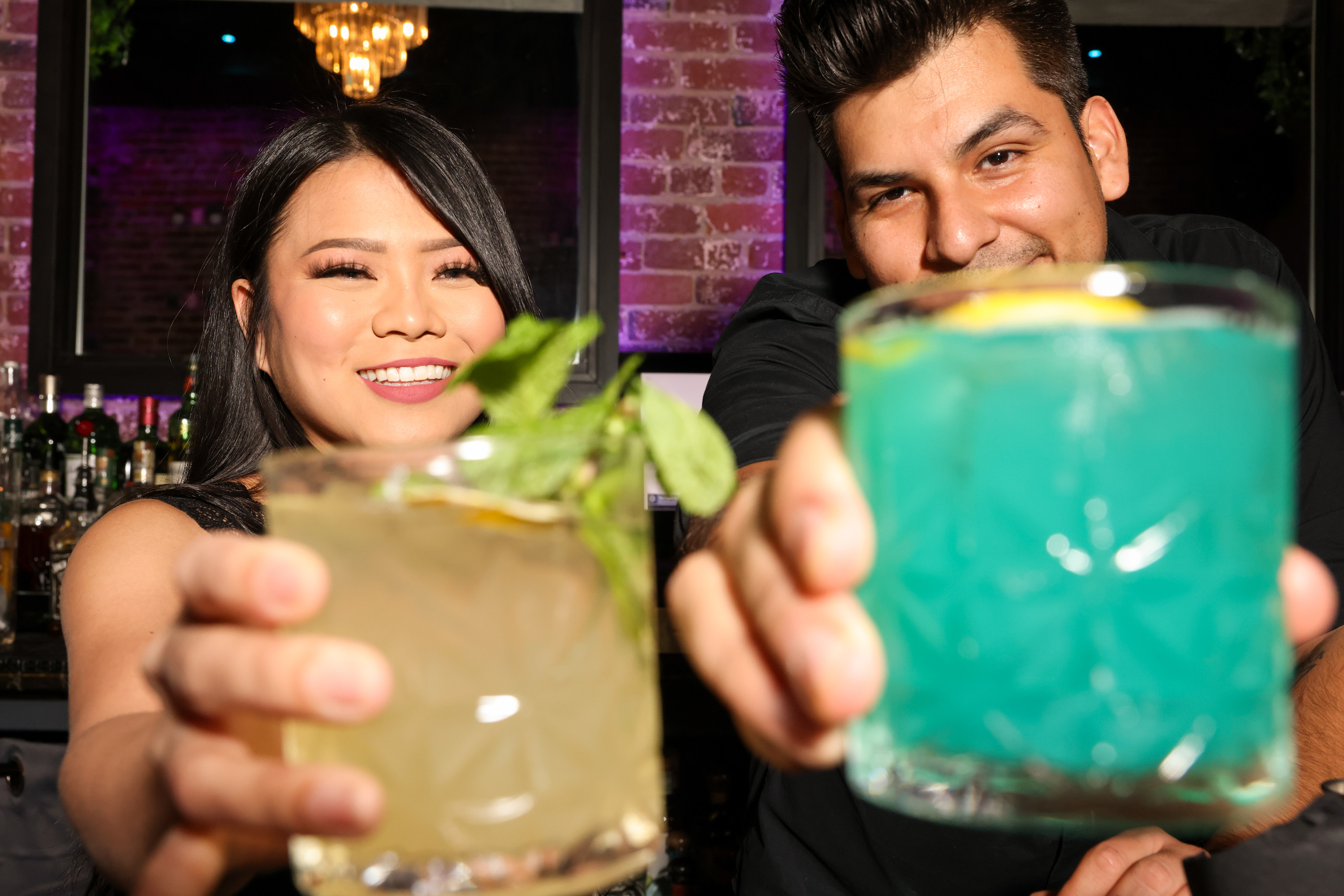 Two smiling people hold colorful cocktails toward the camera, one drink is garnished with mint and the other is blue. They're in a warmly lit bar.