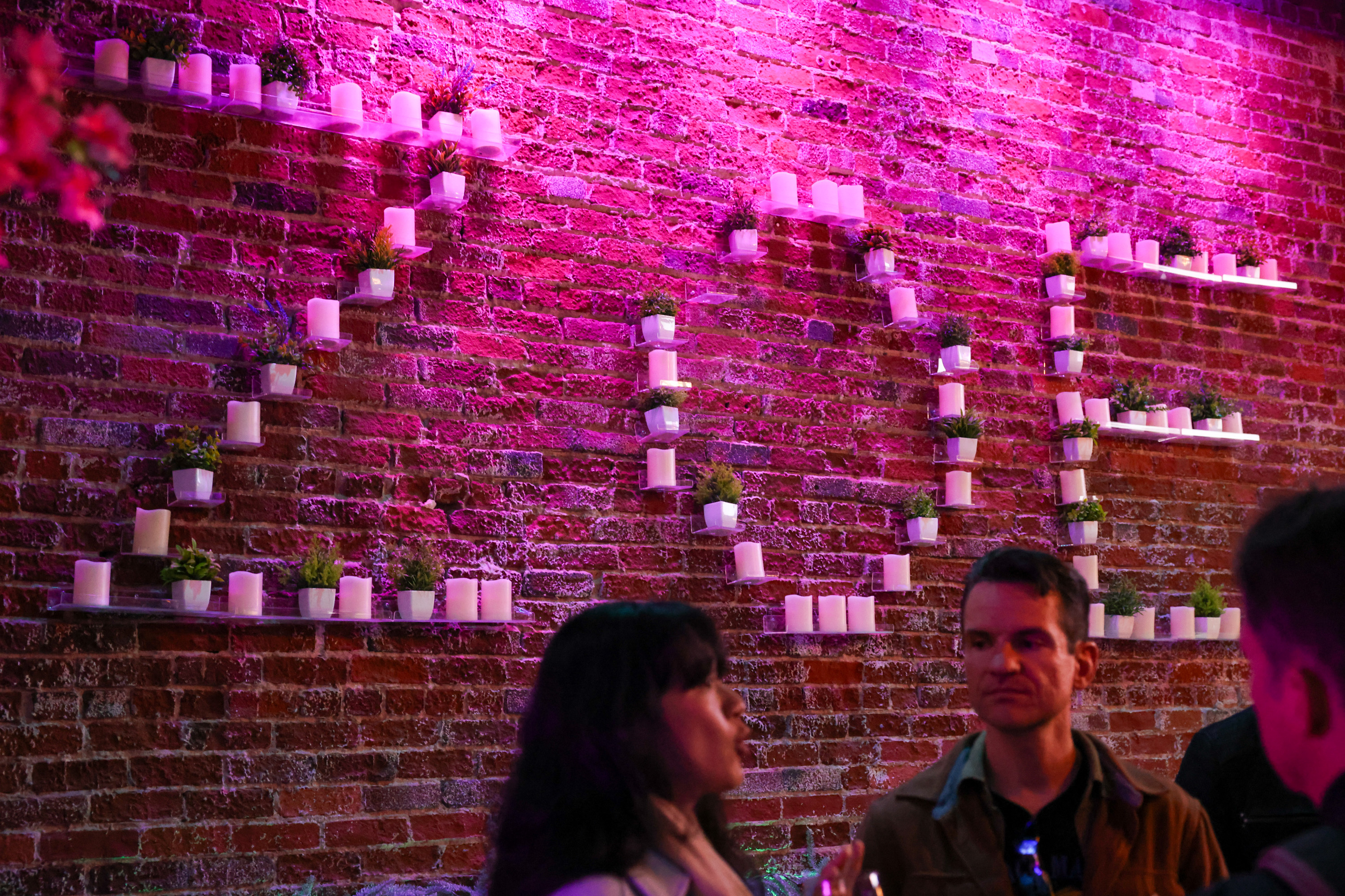 A brick wall is lit with pink lights, decorated with candles and plants forming the numbers &quot;2023.&quot; Two people are conversing in the foreground.