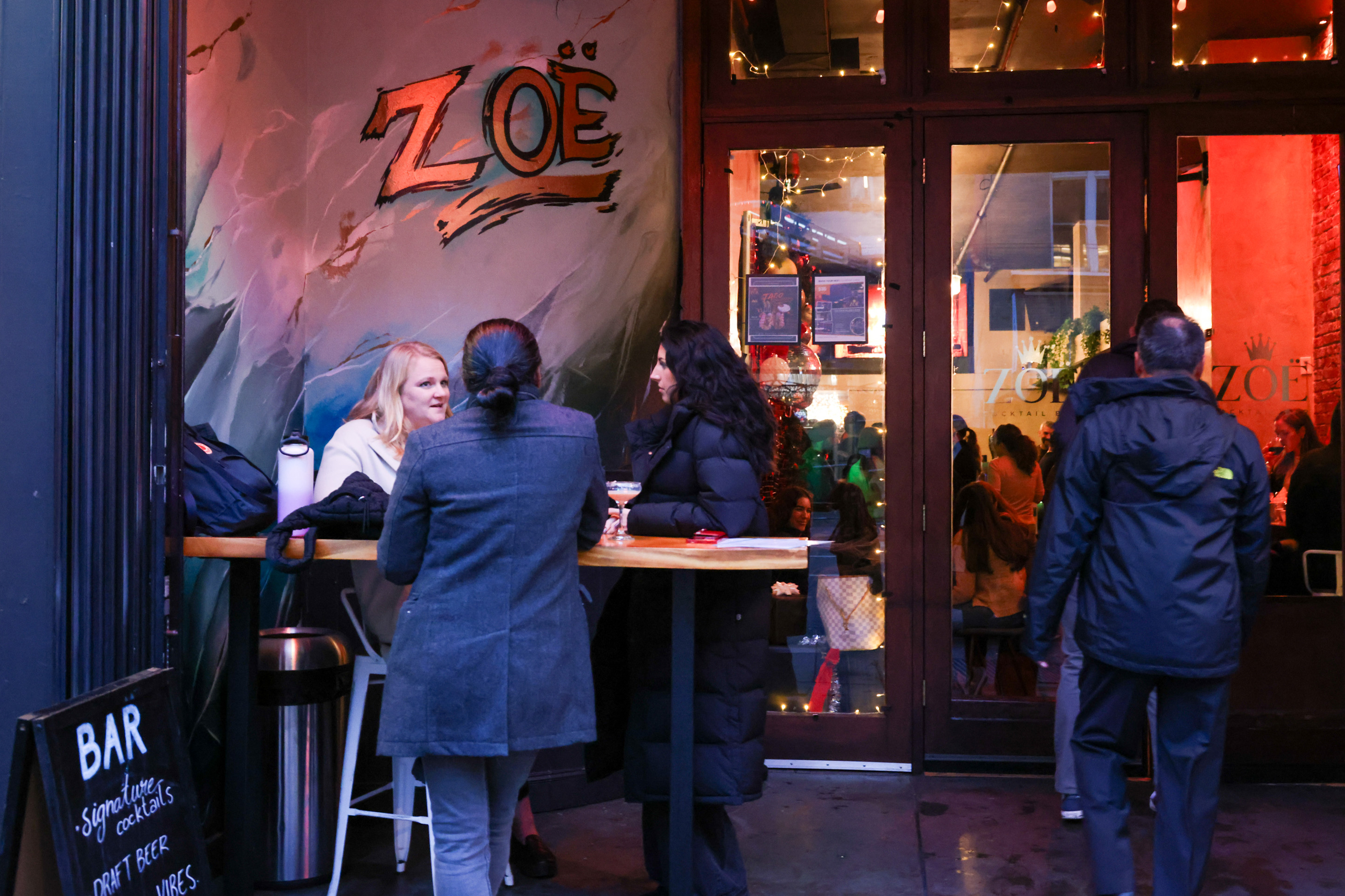 People gathered outside a bar called &quot;Zoe,&quot; chatting around a wooden table. Inside, a lively atmosphere with red lighting and decorations is visible.