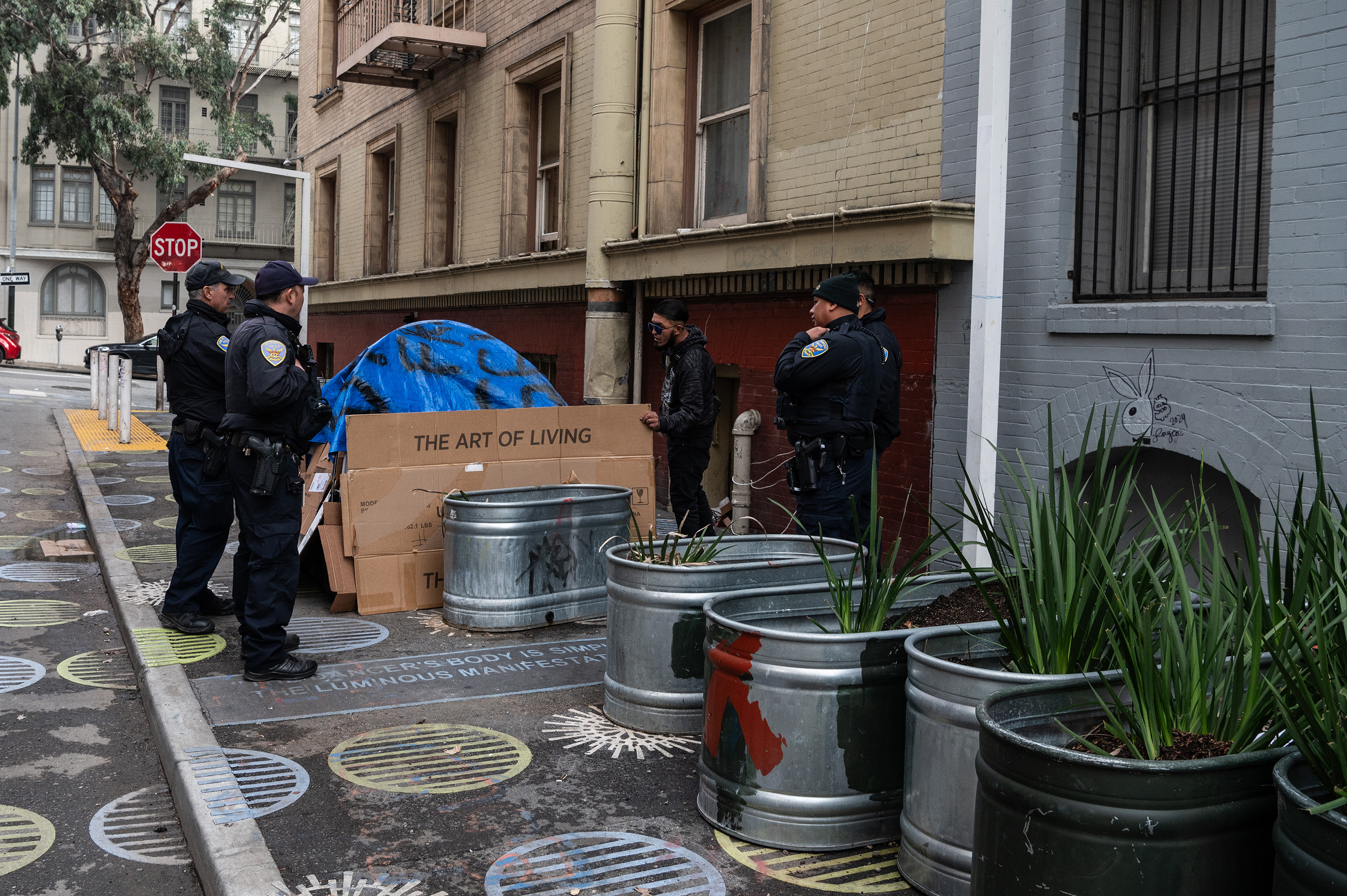 Four police officers are interacting with a man near a building. The man is beside a cardboard shelter labeled &quot;The Art of Living&quot; and some metal planters.