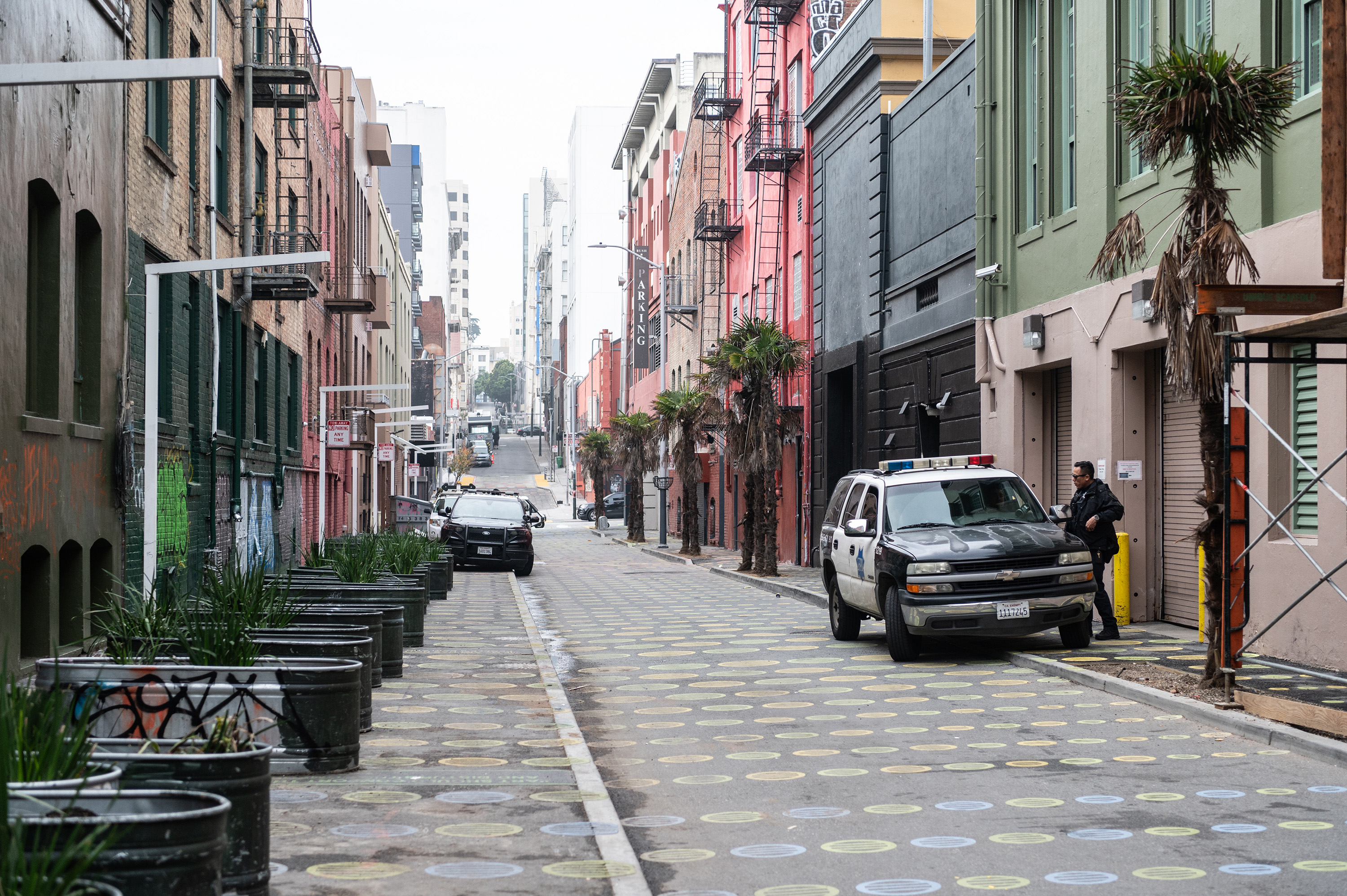 A narrow street with colorful buildings and dotted pavement. Palm trees line the right side, with a police vehicle parked beside them. A person stands nearby.