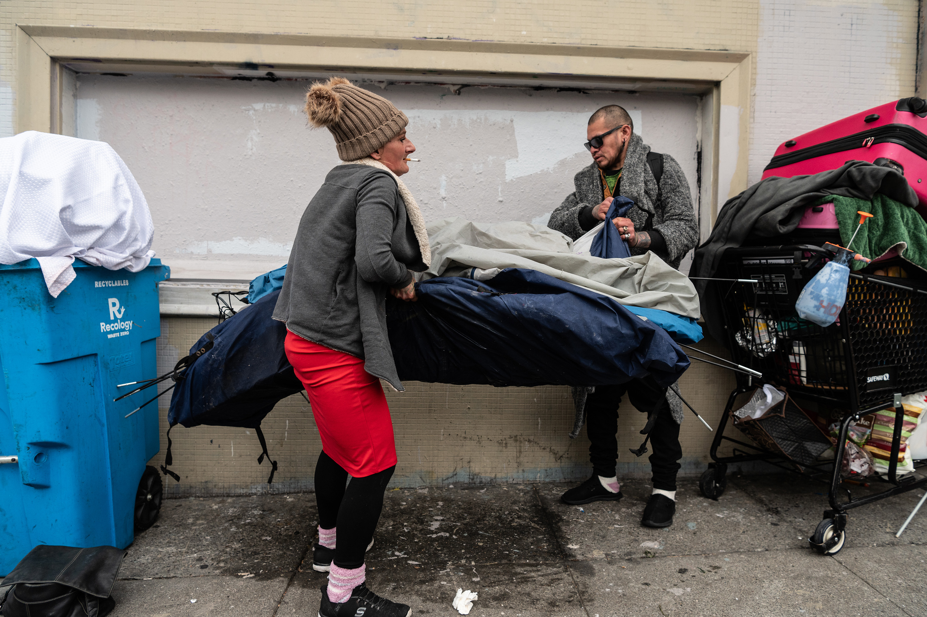 Two people are setting up a tent on a sidewalk. One wears a gray coat and sunglasses, the other a brown hat and red pants. A cart and suitcase are nearby.