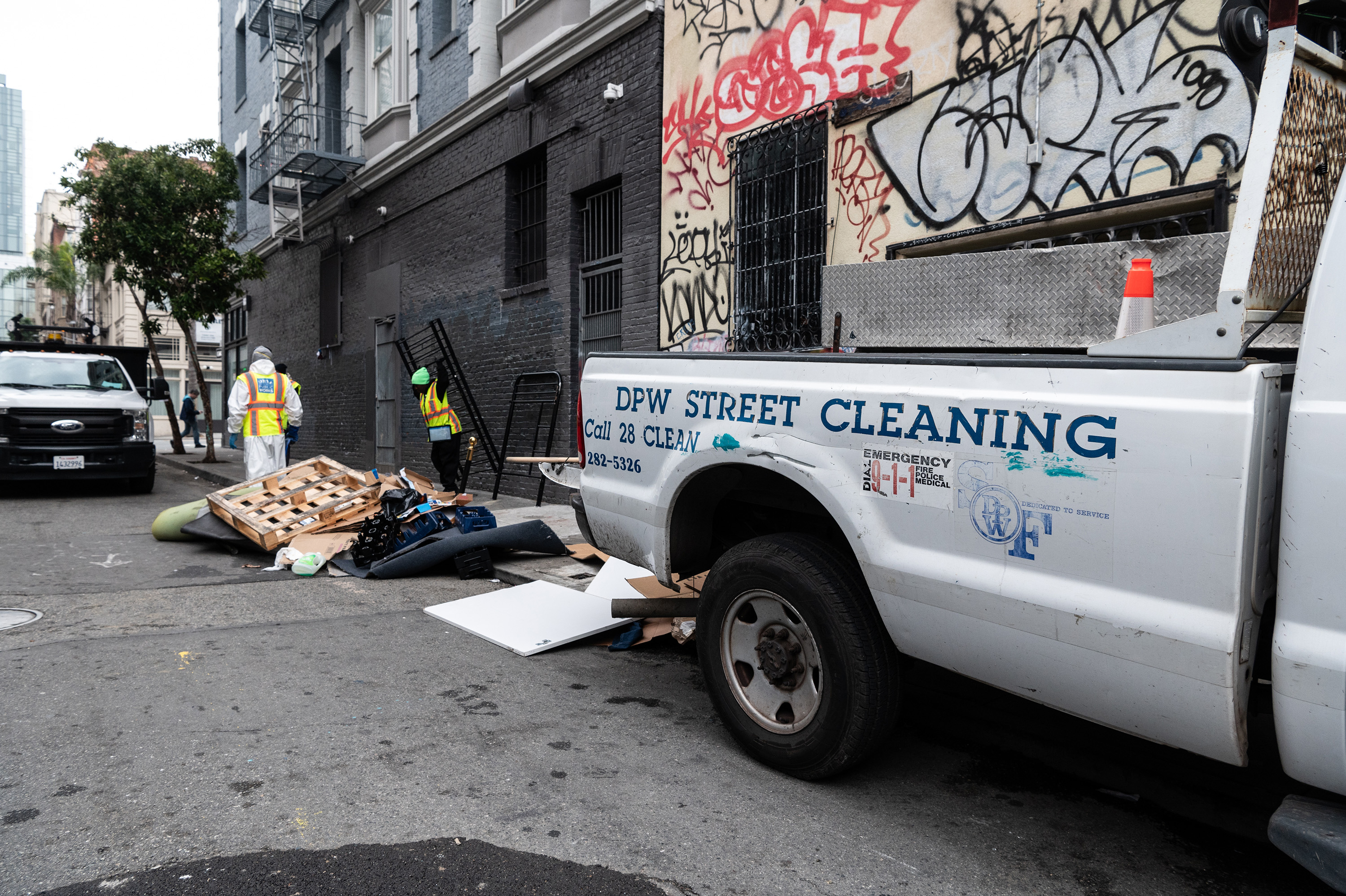 Workers in safety vests clean up debris and wooden pallets beside a graffiti-covered wall. A DPW street cleaning truck is parked on the street.