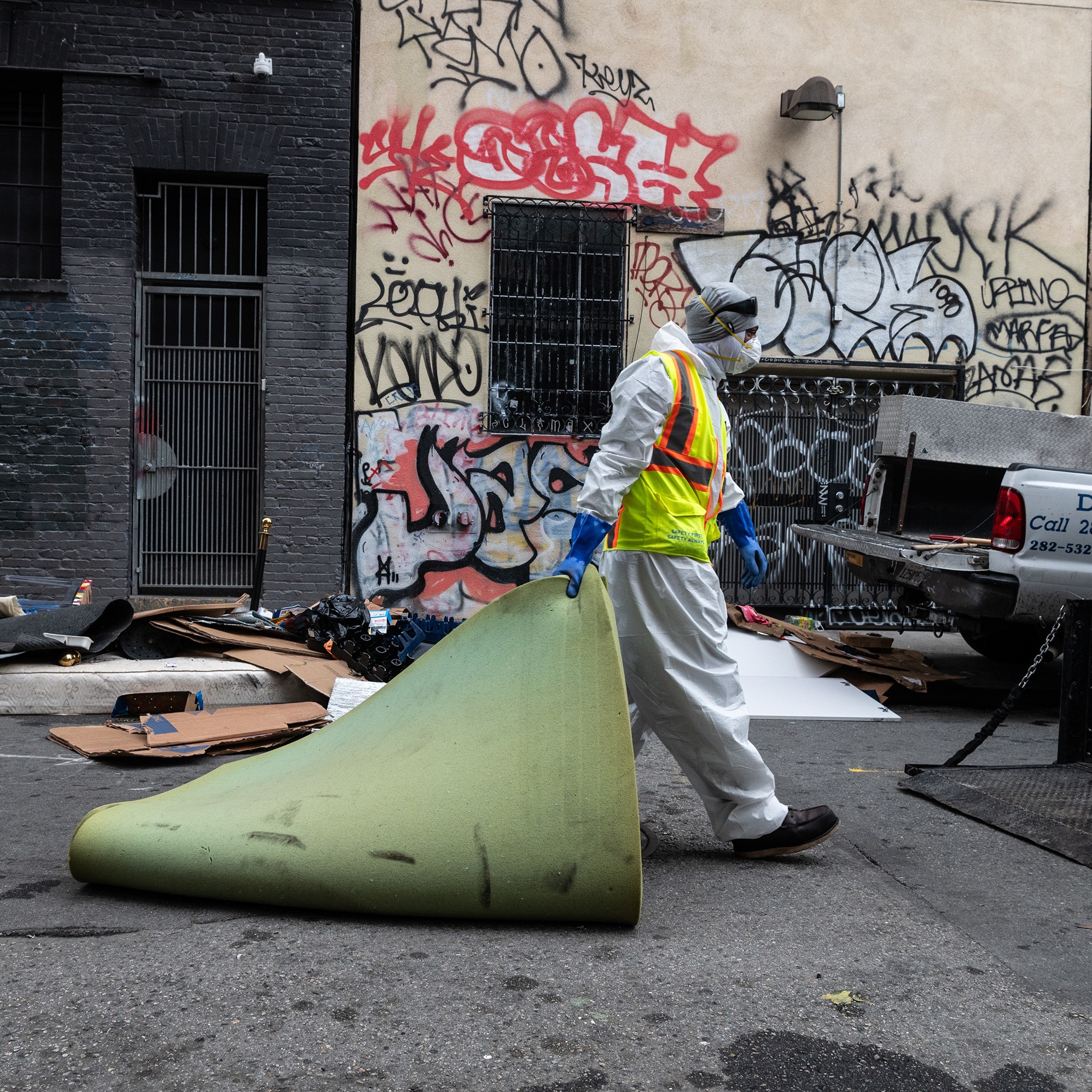 A person in protective gear carries a rolled-up carpet in an alley with graffiti-covered walls. Debris and a truck labeled "DPW" are visible around.