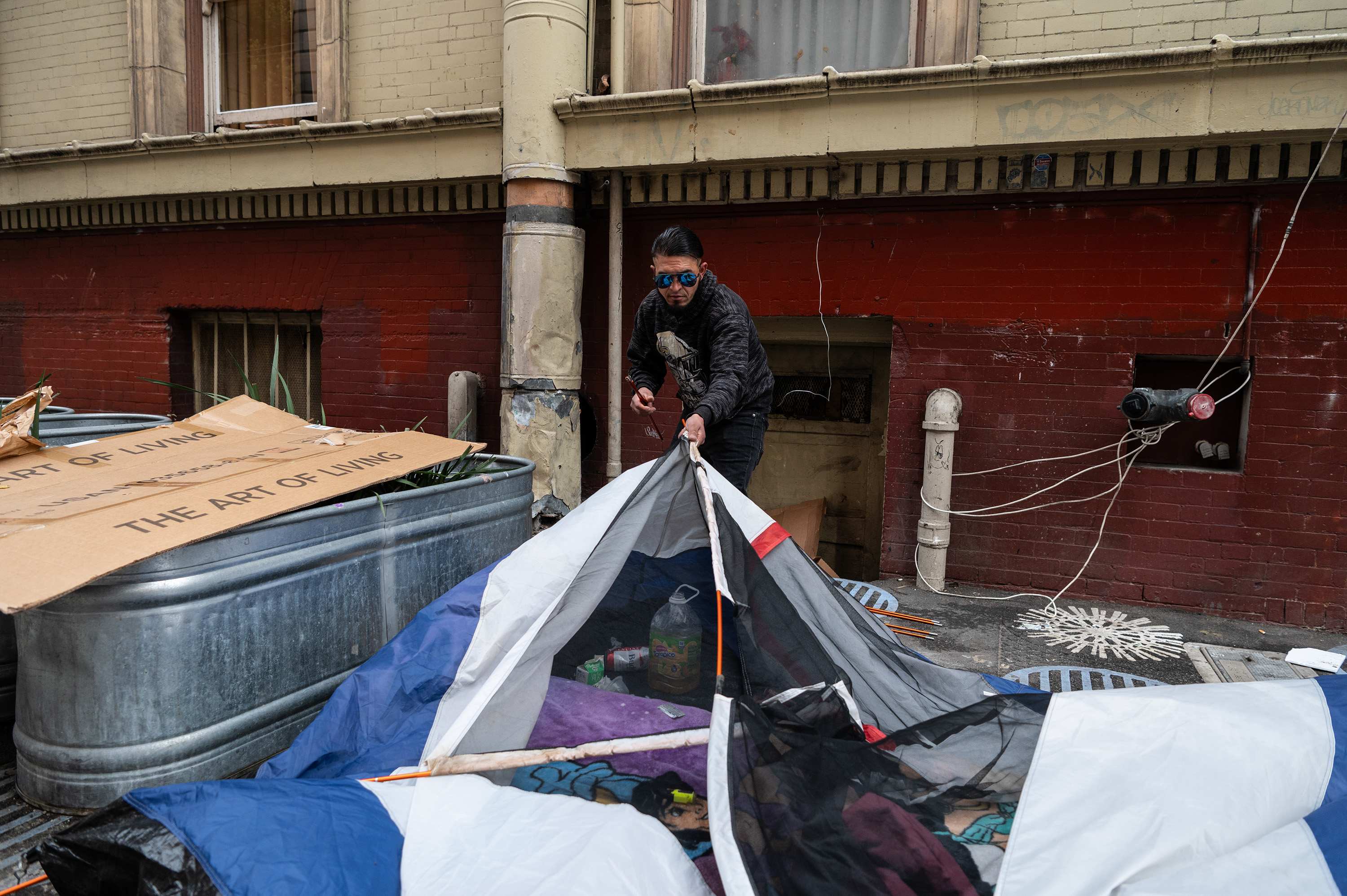 A person in sunglasses is setting up a multi-colored tent on a city street, surrounded by cardboard and metal containers, against a brick wall.