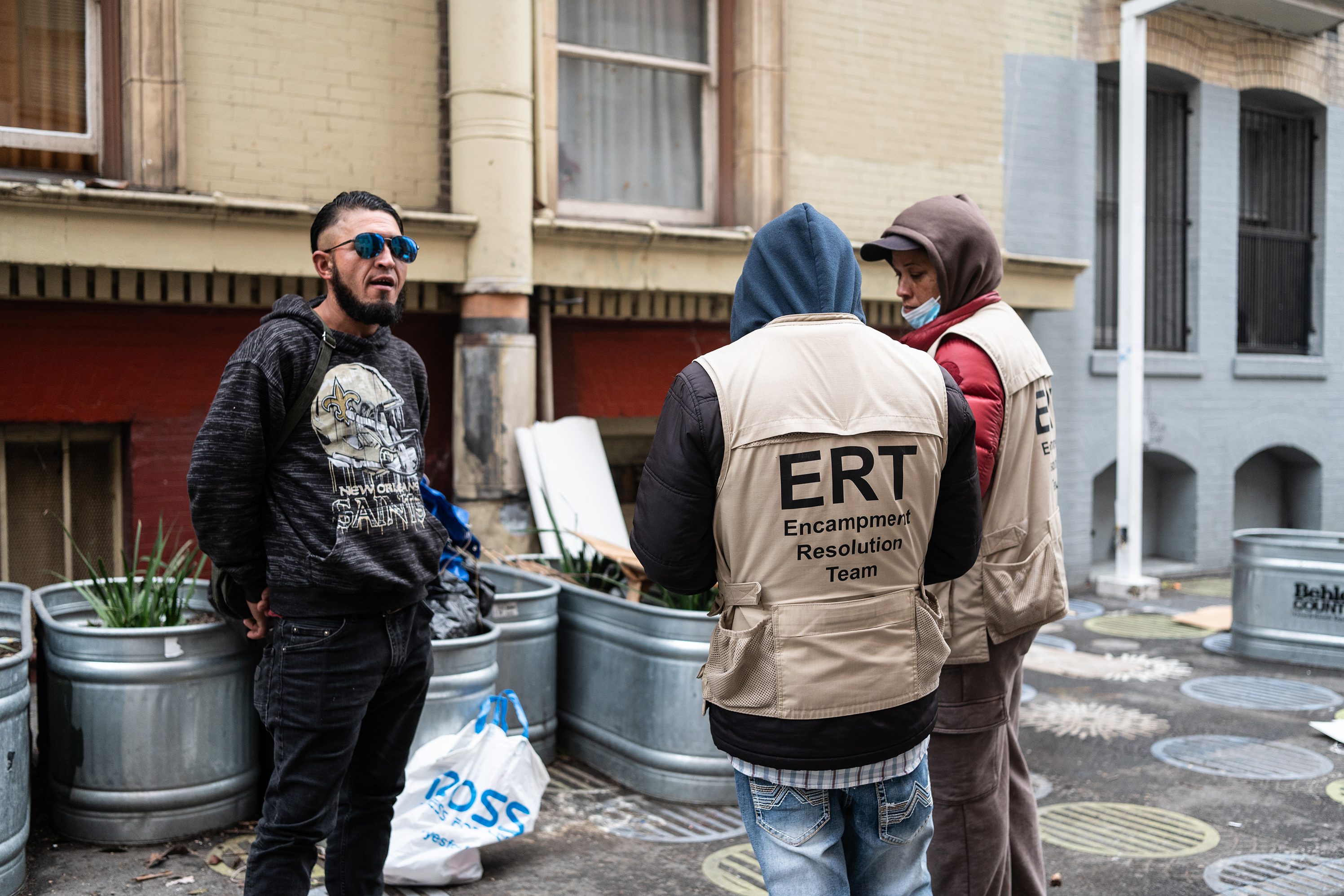 Three people are talking outside. One wears sunglasses and a dark hoodie, while the other two wear &quot;ERT Encampment Resolution Team&quot; vests.