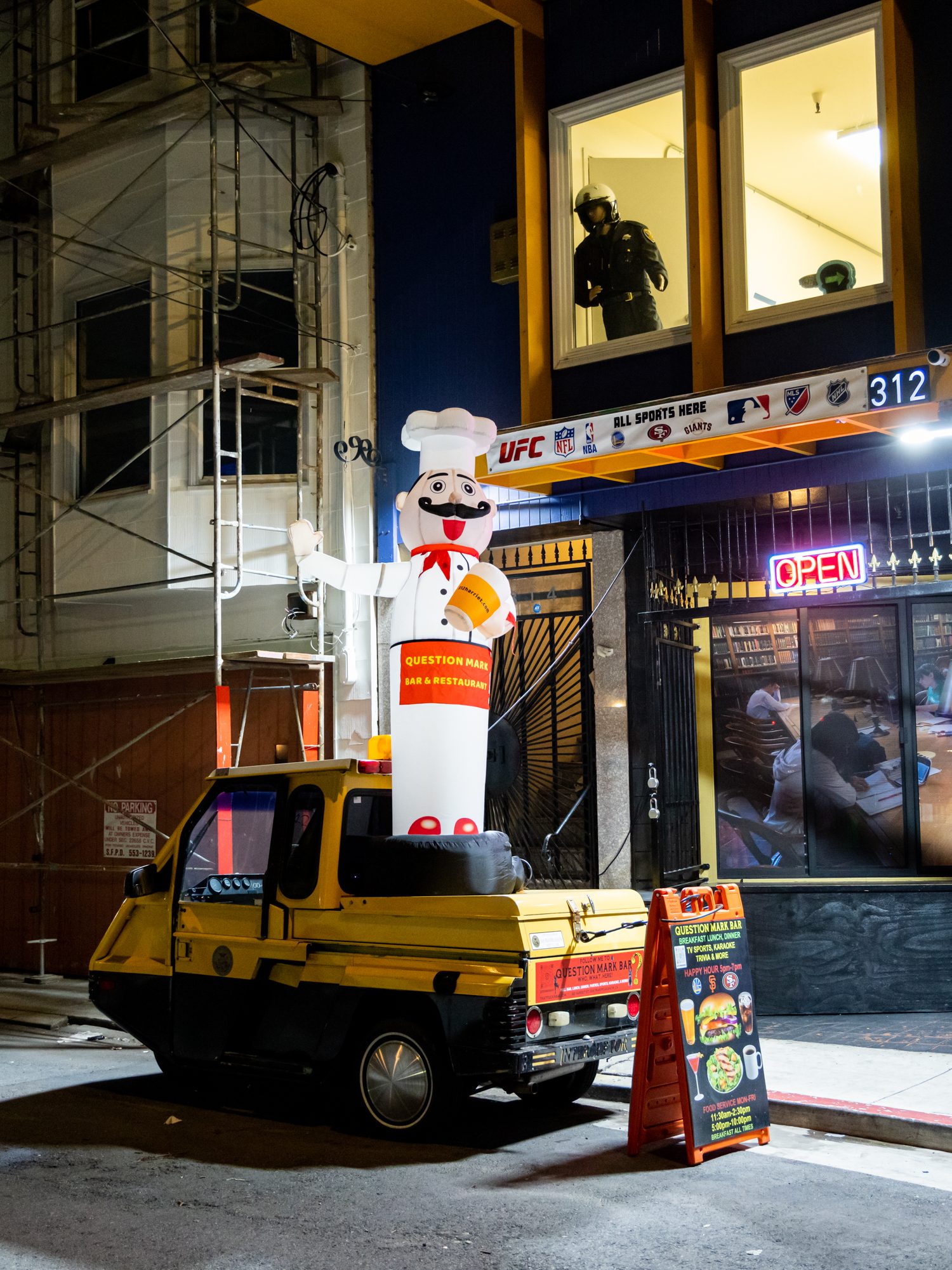 A small yellow vehicle is parked by a restaurant. A large inflatable chef stands beside it. Above, a window displays a mannequin wearing a helmet.