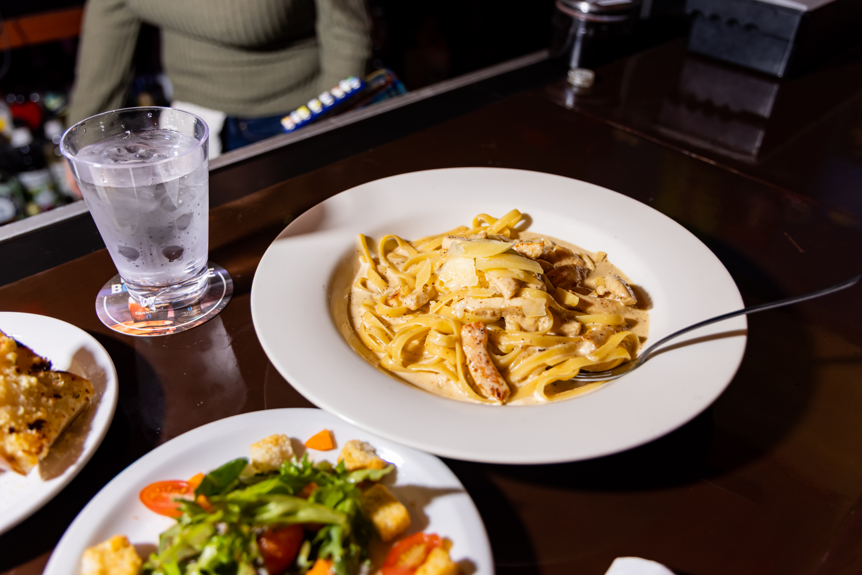 A plate of creamy pasta with chicken and parmesan sits on a dark table, next to a fresh salad, garlic bread, and a glass of ice water.