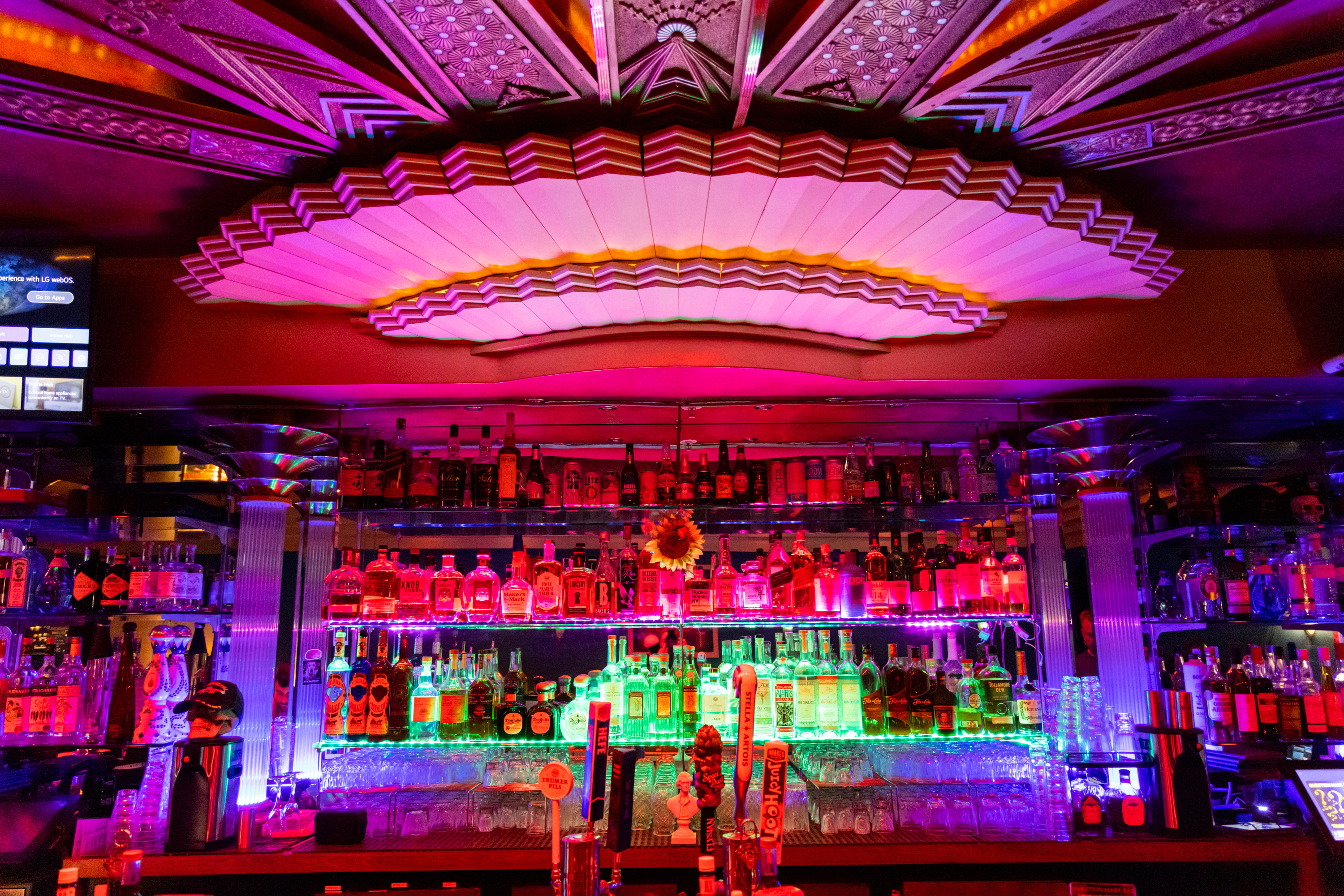 A vibrant bar top with colorful bottles backlit in pink and green. There's an ornate ceiling with geometric designs and decorative columns.