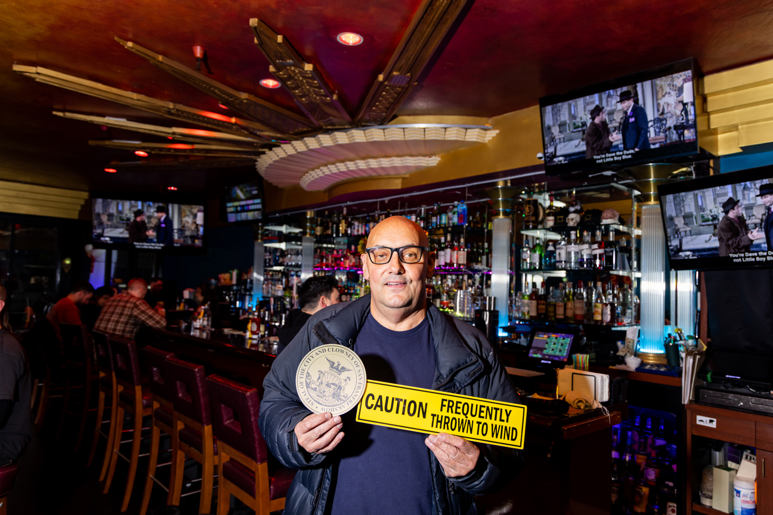 A man in a bar holds a round plaque and a yellow sign reading &quot;Caution Frequently Thrown to Wind&quot; with patrons seated at the bar and TVs in the background.