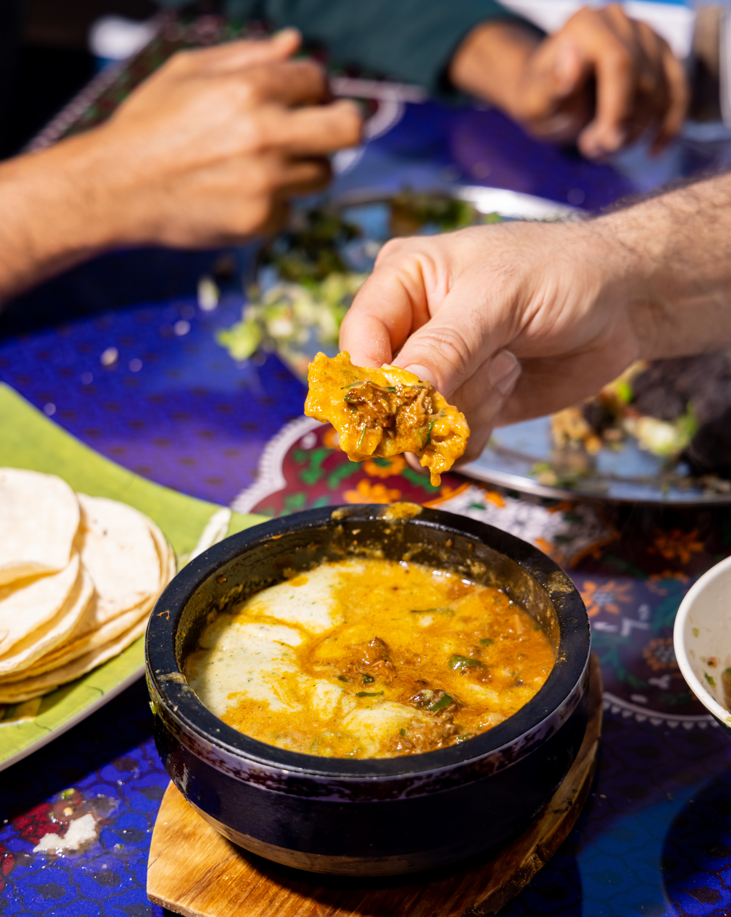 A hand dips bread into a creamy curry in a black bowl. There are tortillas on a plate nearby, and various dishes on a colorful tablecloth in the background.