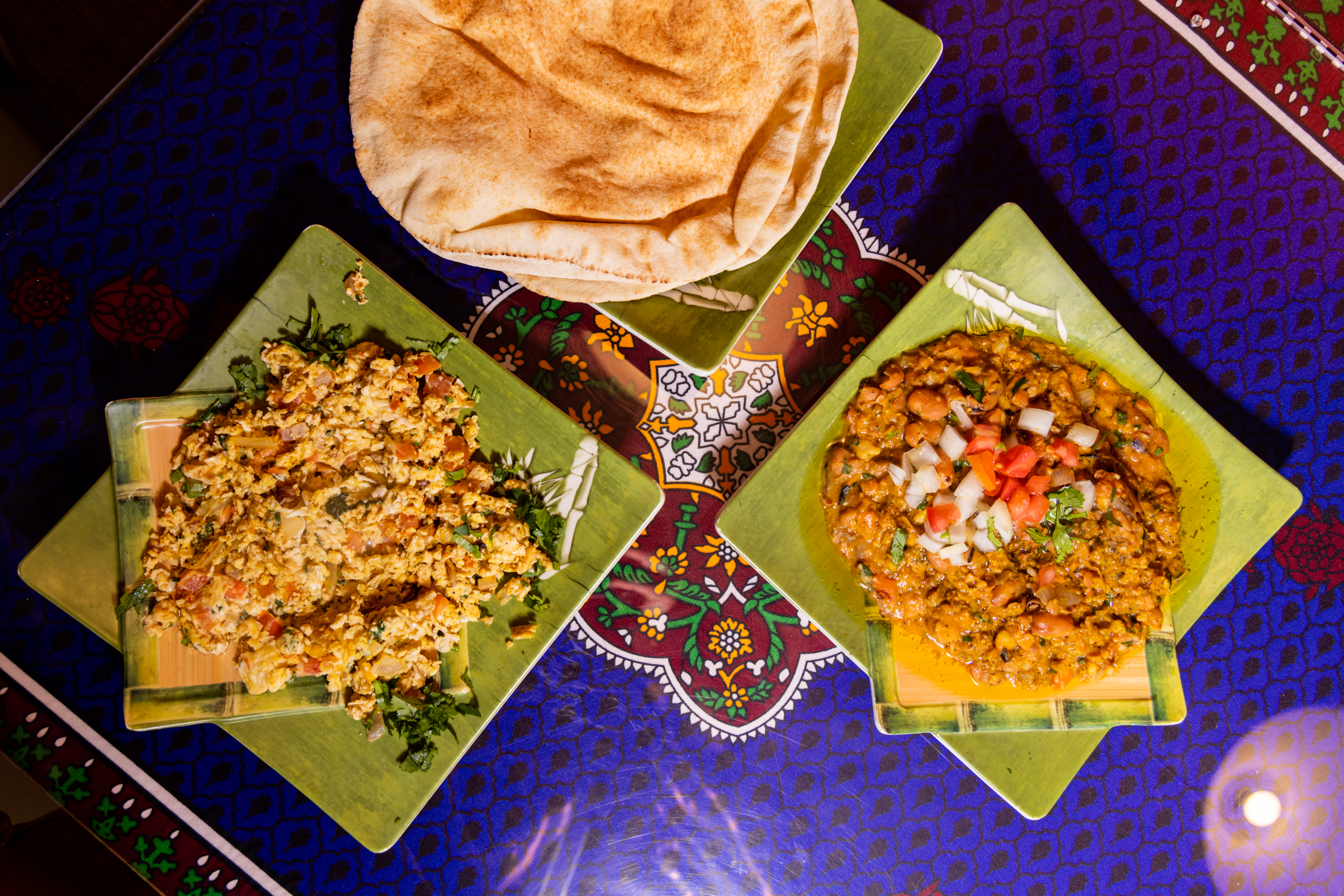 Three green plates are on a colorful tablecloth: one with a scrambled mixture, another with a vegetable stew garnished with diced onions and tomatoes, and a plate of flatbread.