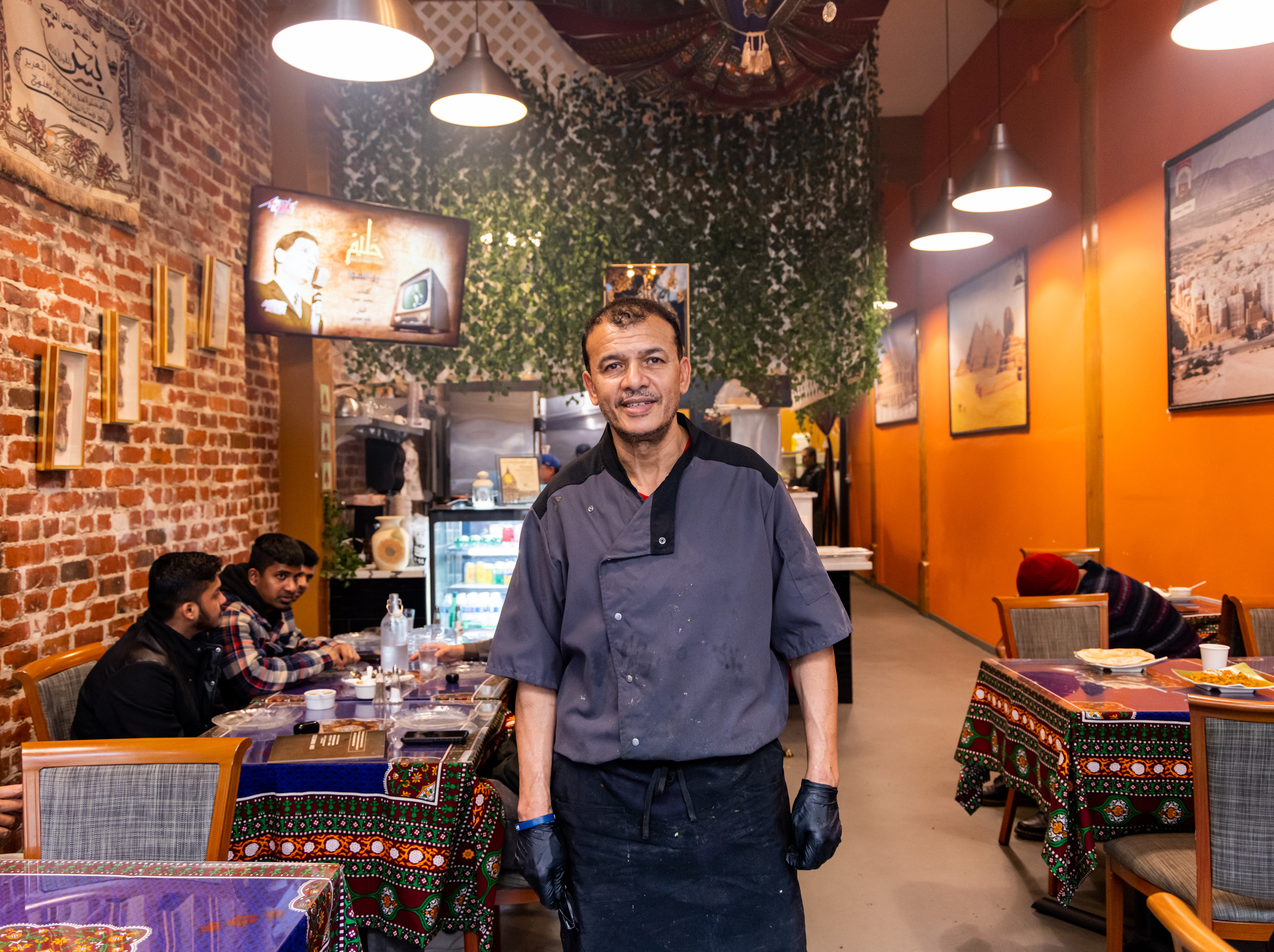 A chef wearing a black uniform stands smiling inside a cozy restaurant with brick walls and hanging lights. Patrons are seated at tables covered in colorful cloth.