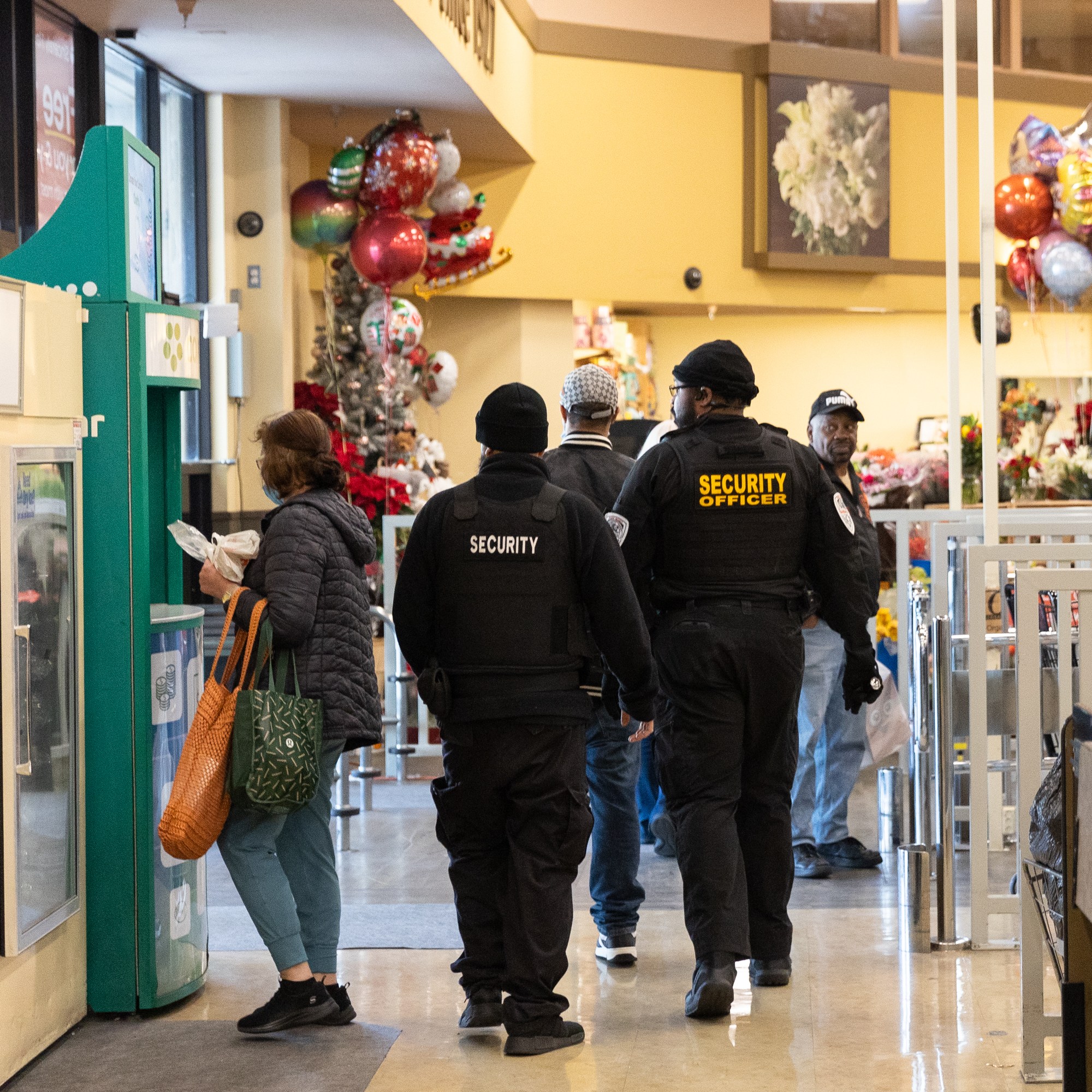 Security personnel in black uniforms walk through a brightly lit grocery store near a floral display and balloons, while a customer stands nearby.