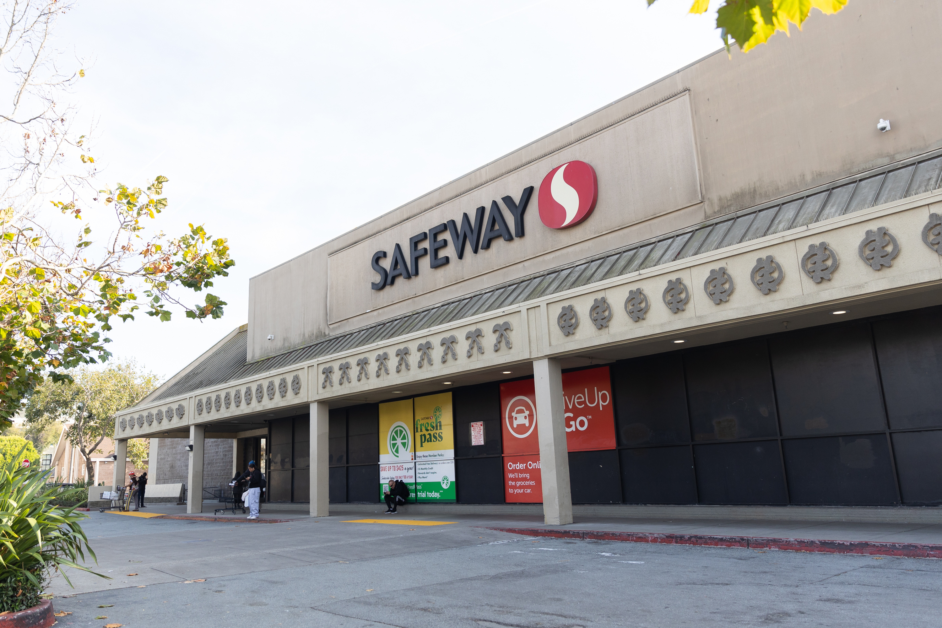 The image shows the exterior of a Safeway supermarket with a wide entrance, decorated roof, and signs for services. A tree with green leaves is visible on the left.