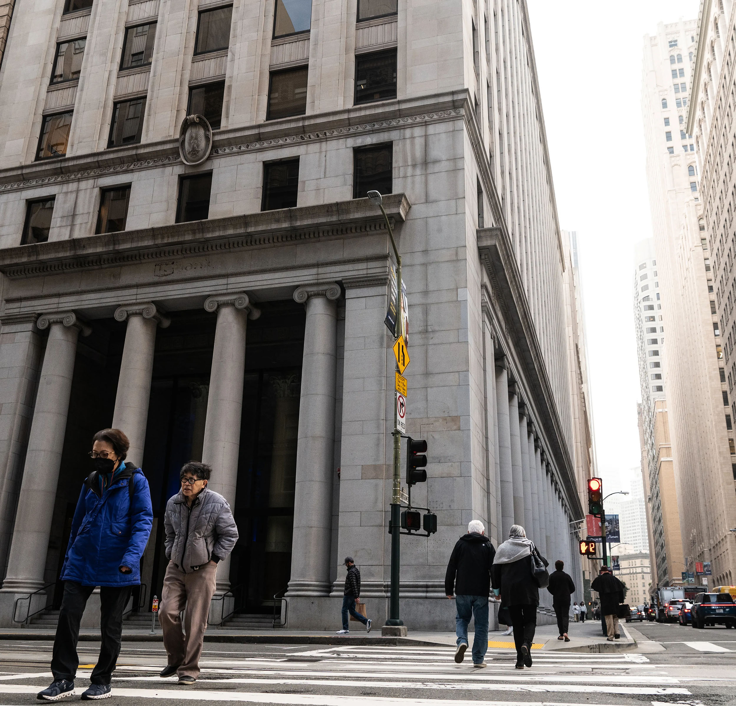 People cross a street in front of a tall, stone building with large columns. The city scene is busy, with others walking and a few cars visible in the distance.