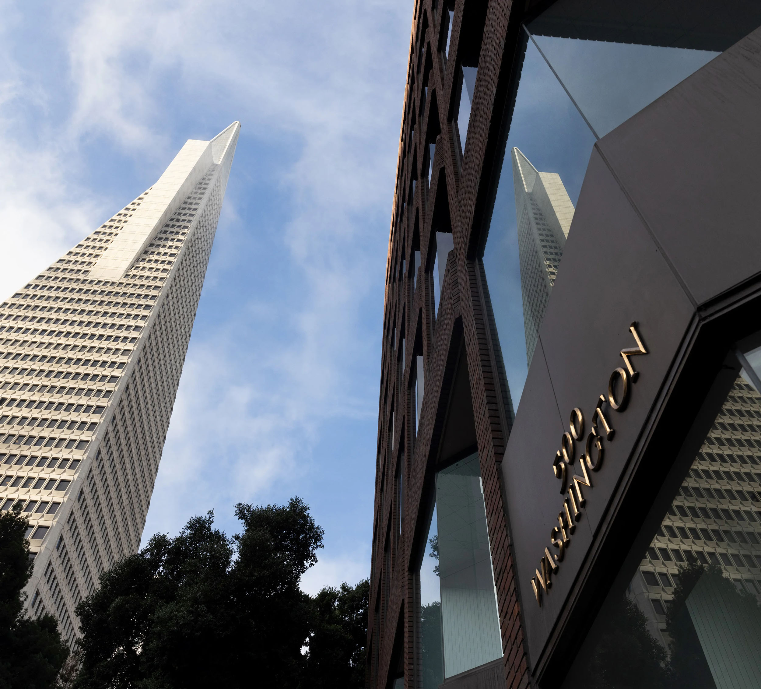 A tall, pointed skyscraper rises against a blue sky next to a building with the address &quot;600 Washington&quot; and reflects in its windows. Trees are visible below.