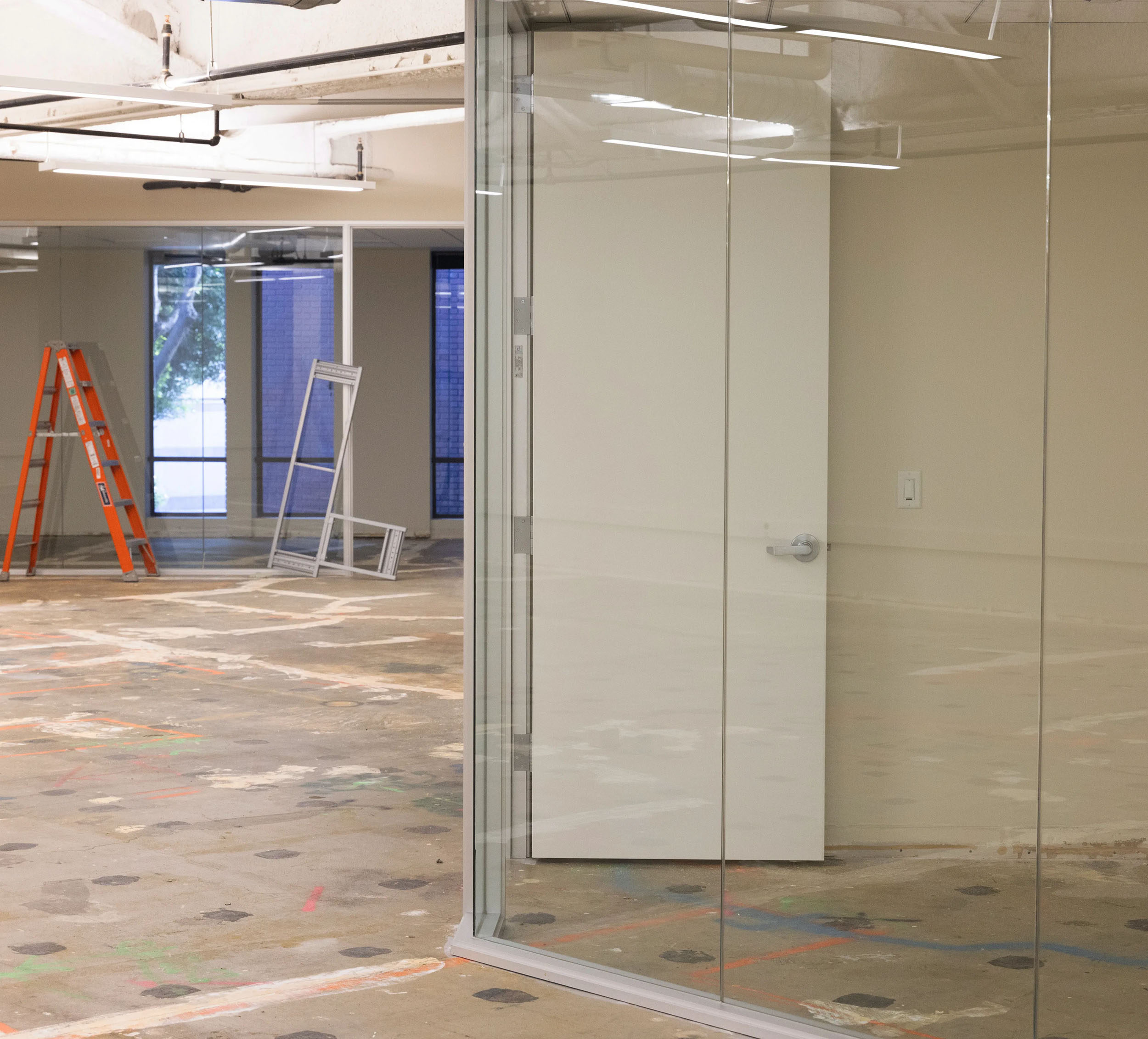 The image shows an unfinished office space with glass walls, an open white door, exposed flooring, an orange ladder, and window frames propped against the wall.