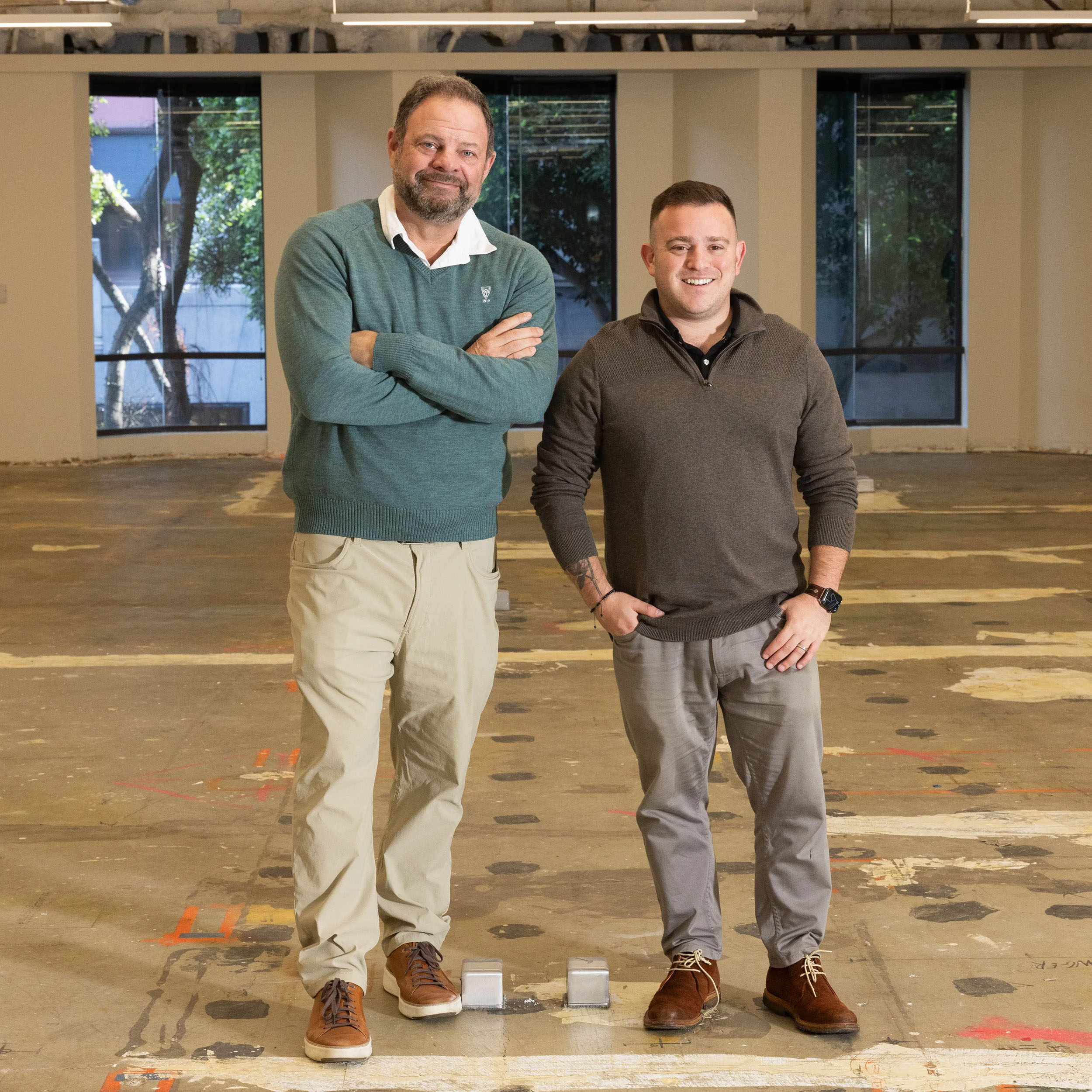 Two men stand in a large, unfinished room with a concrete floor. The man on the left wears a green sweater, and the man on the right wears a gray sweater.