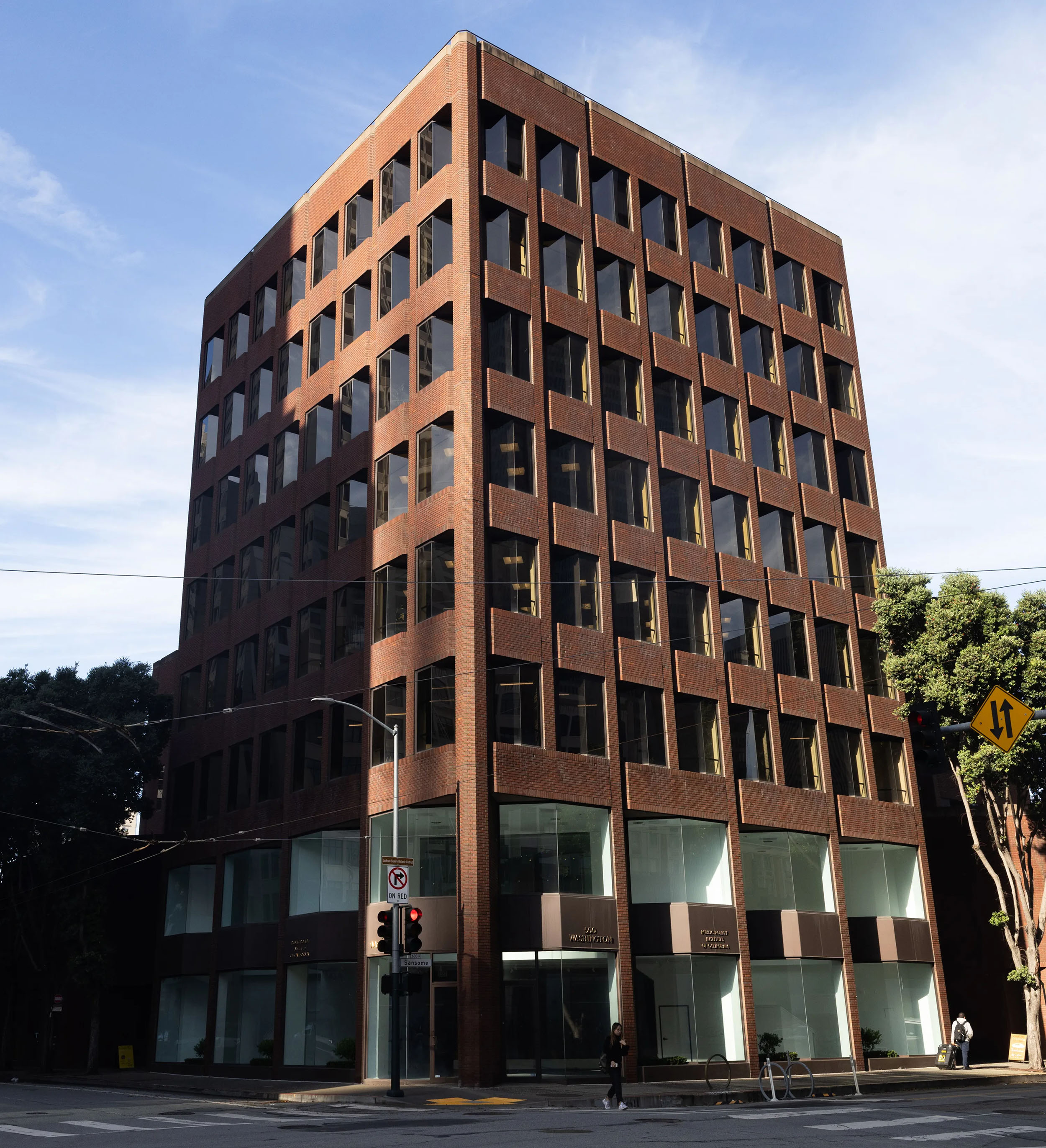 A multi-story brick office building stands on a street corner under a clear sky. It has large windows and a modern design. A few trees and street signs are visible.