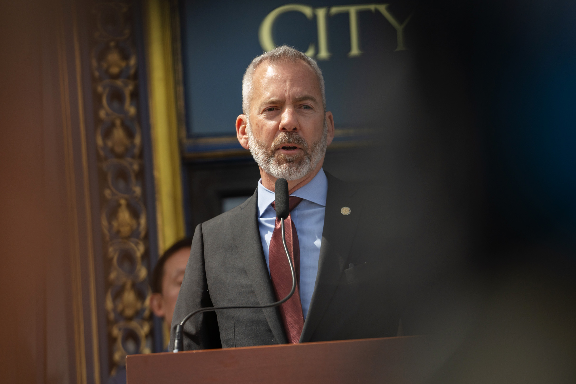 A man with a beard in a suit and red tie speaks at a podium with a microphone, in front of a decorative background. Part of the word "CITY" is visible.