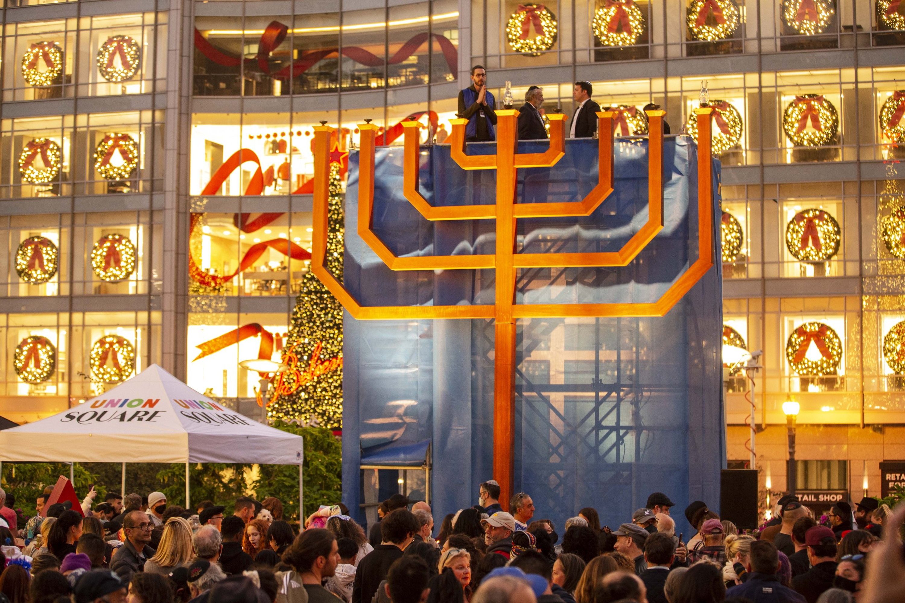 A large menorah is lit in a festive outdoor gathering. People crowd around, and a decorated building with wreaths and ribbons is visible in the background.