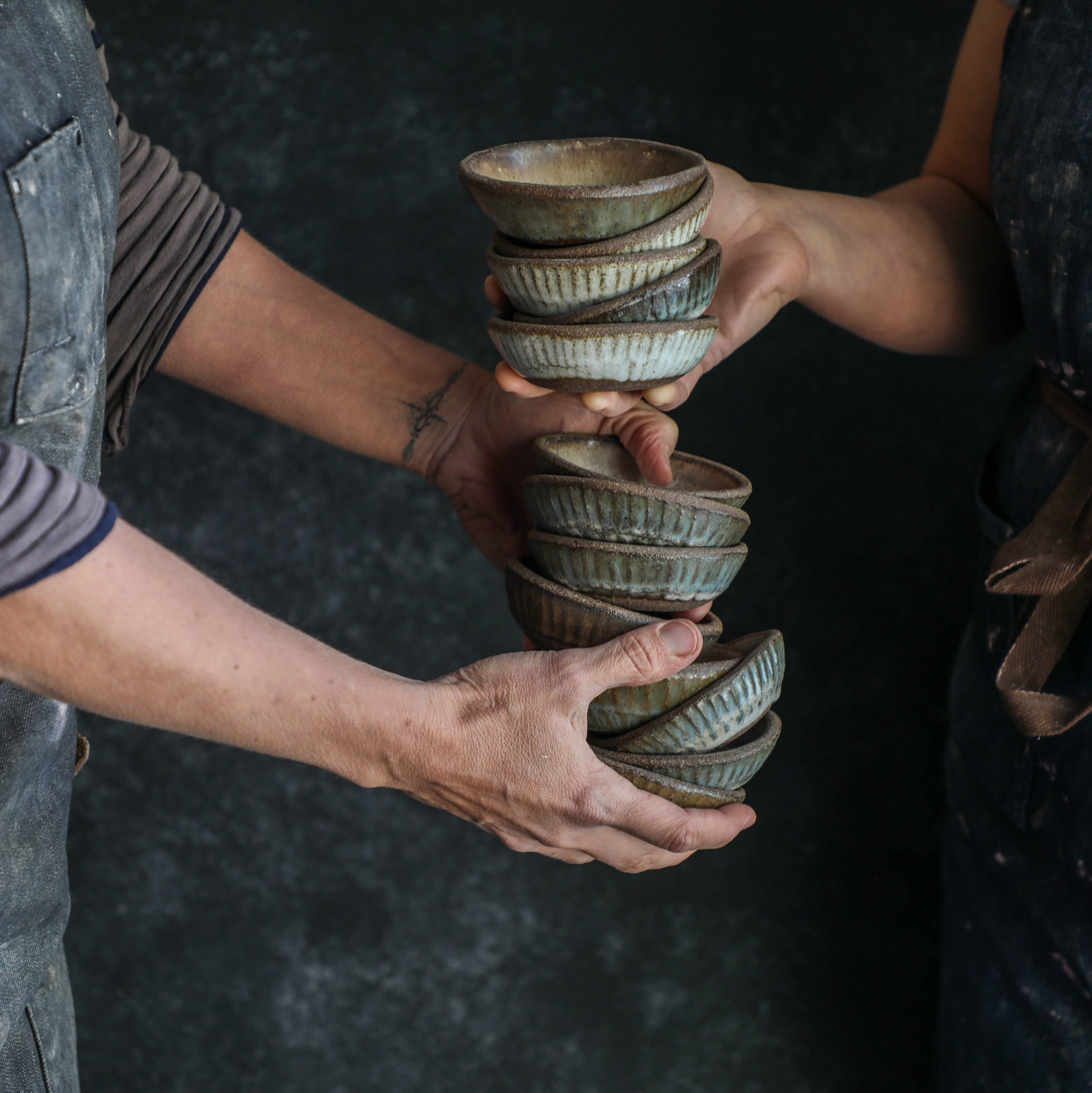 Two people in aprons are exchanging a stack of small, textured ceramic bowls against a dark background.