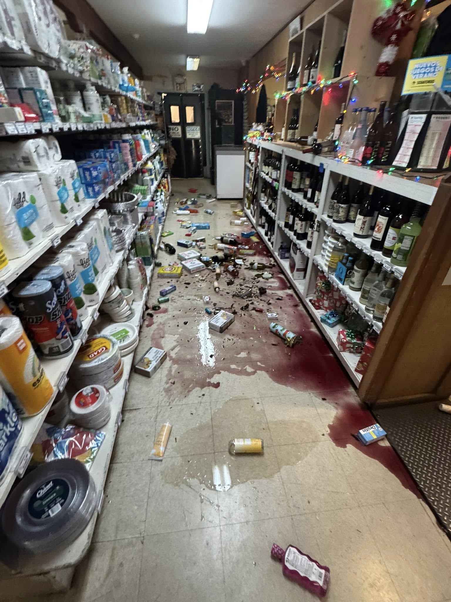 A supermarket aisle is in disarray, with broken bottles and spilled liquids, possibly wine, covering the floor. Shelves are stocked with groceries and festive lights.