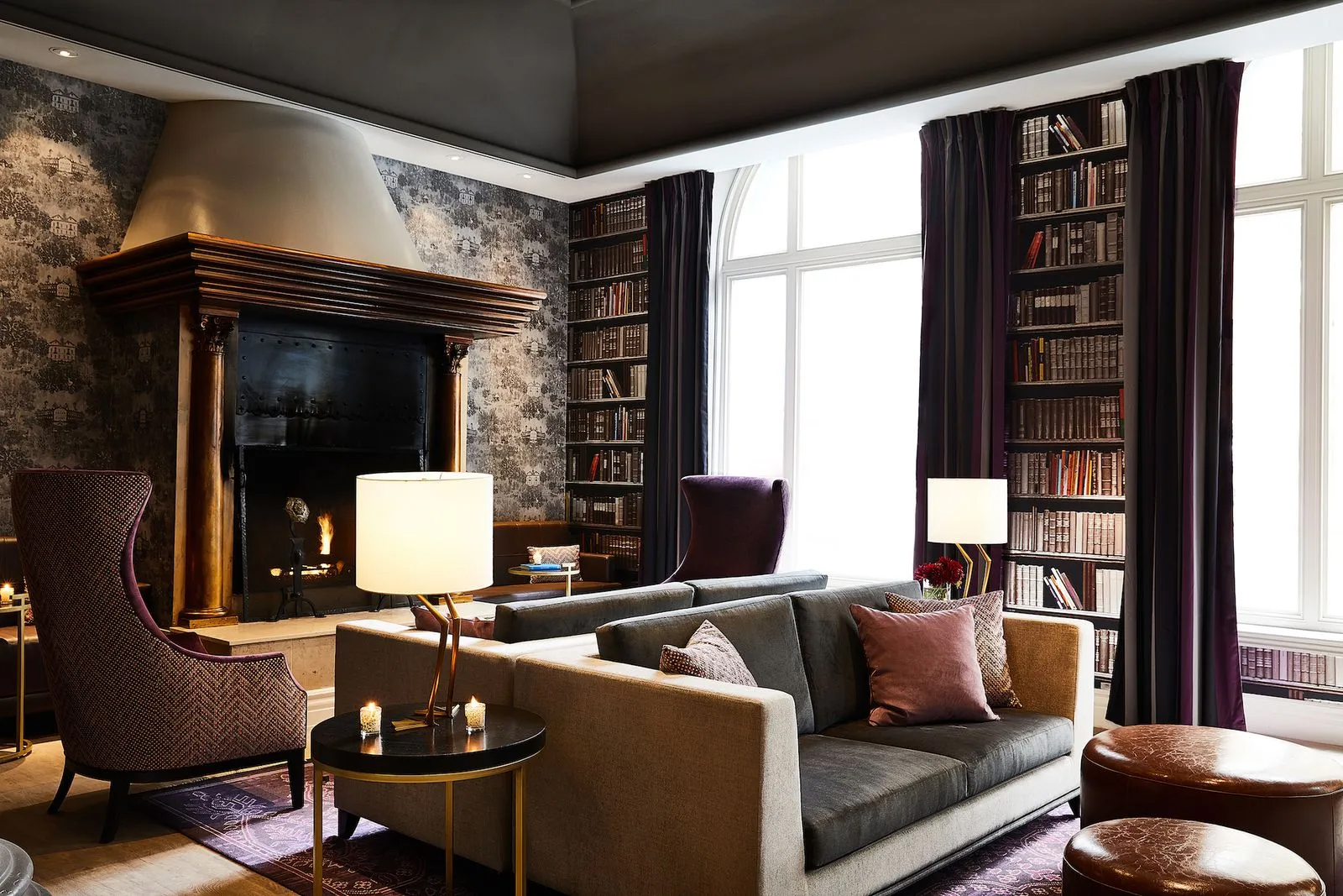 A cozy room features a large stone fireplace, bookshelves, plush seating, and soft lighting. Dark curtains frame tall windows, creating a warm, inviting atmosphere.