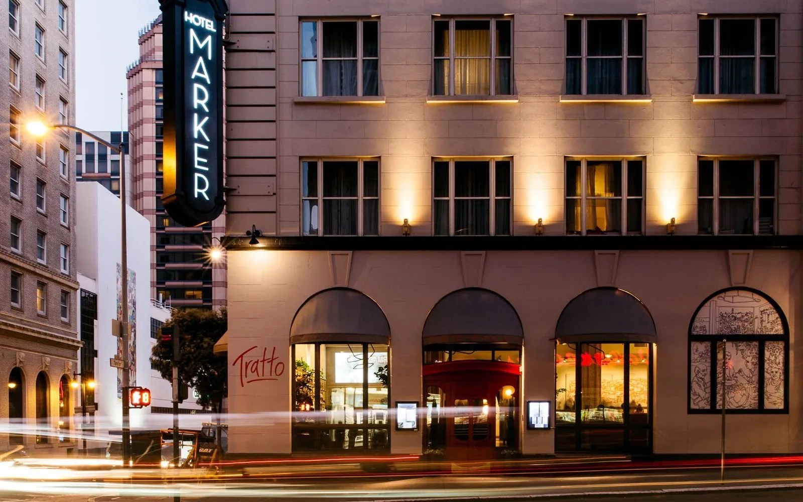 The image shows the exterior of Hotel Marker at dusk, with lit windows and a neon sign. Below, &quot;Tratto&quot; is visible near arched windows, and car lights stream by.