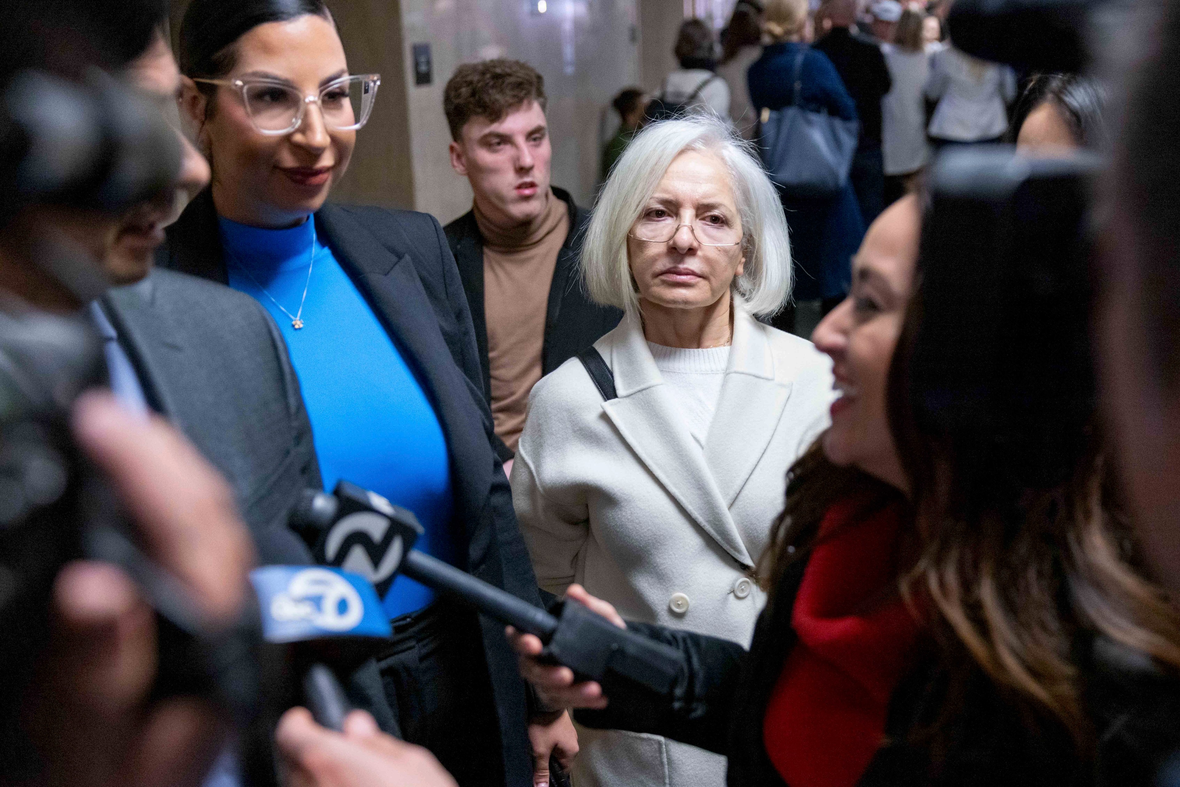 In a crowded hallway, a woman wearing glasses and a white coat looks serious, while microphones are pointed toward her and others in the foreground.