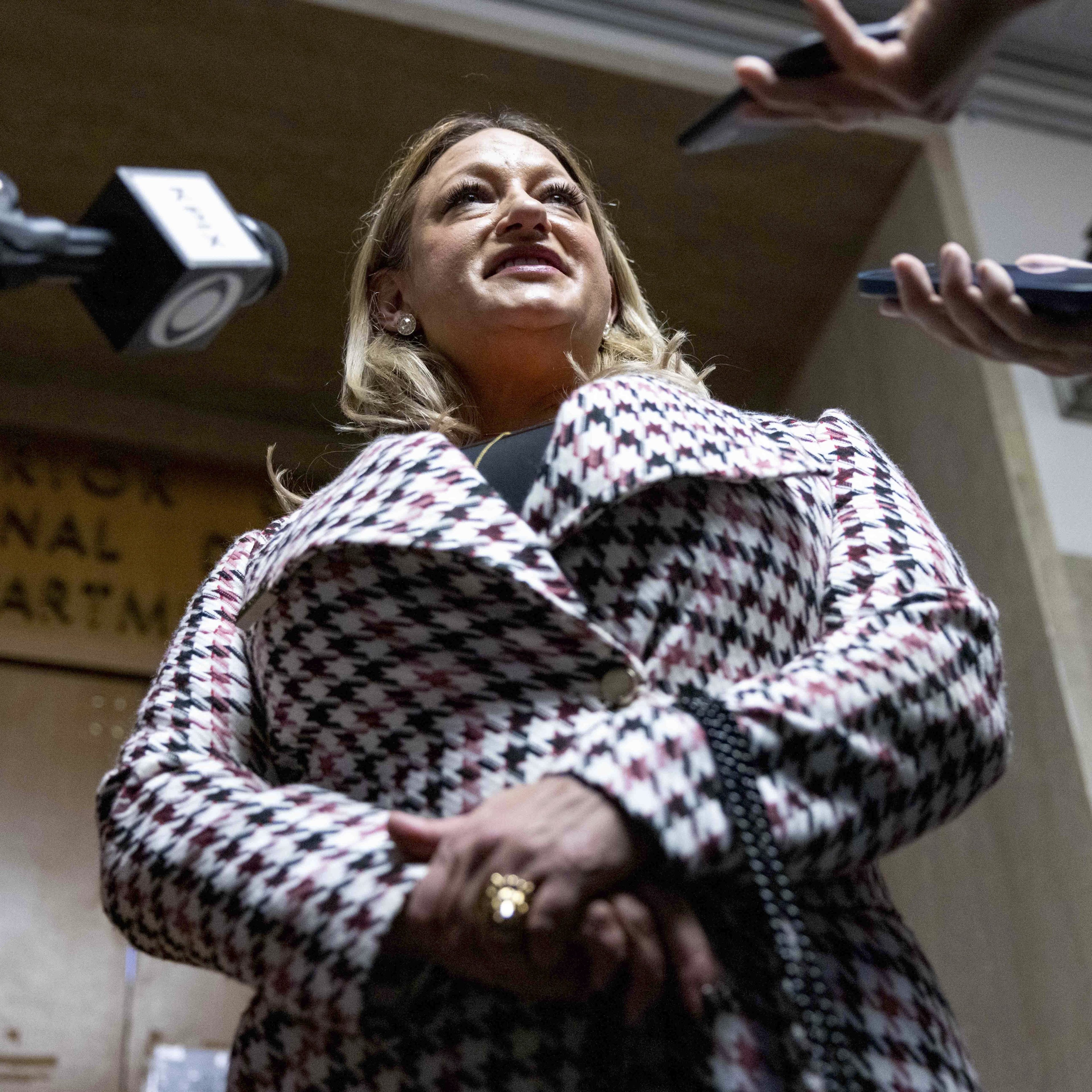 A woman in a houndstooth coat is speaking to multiple reporters holding microphones and phones, standing in front of a wooden door with an official sign.