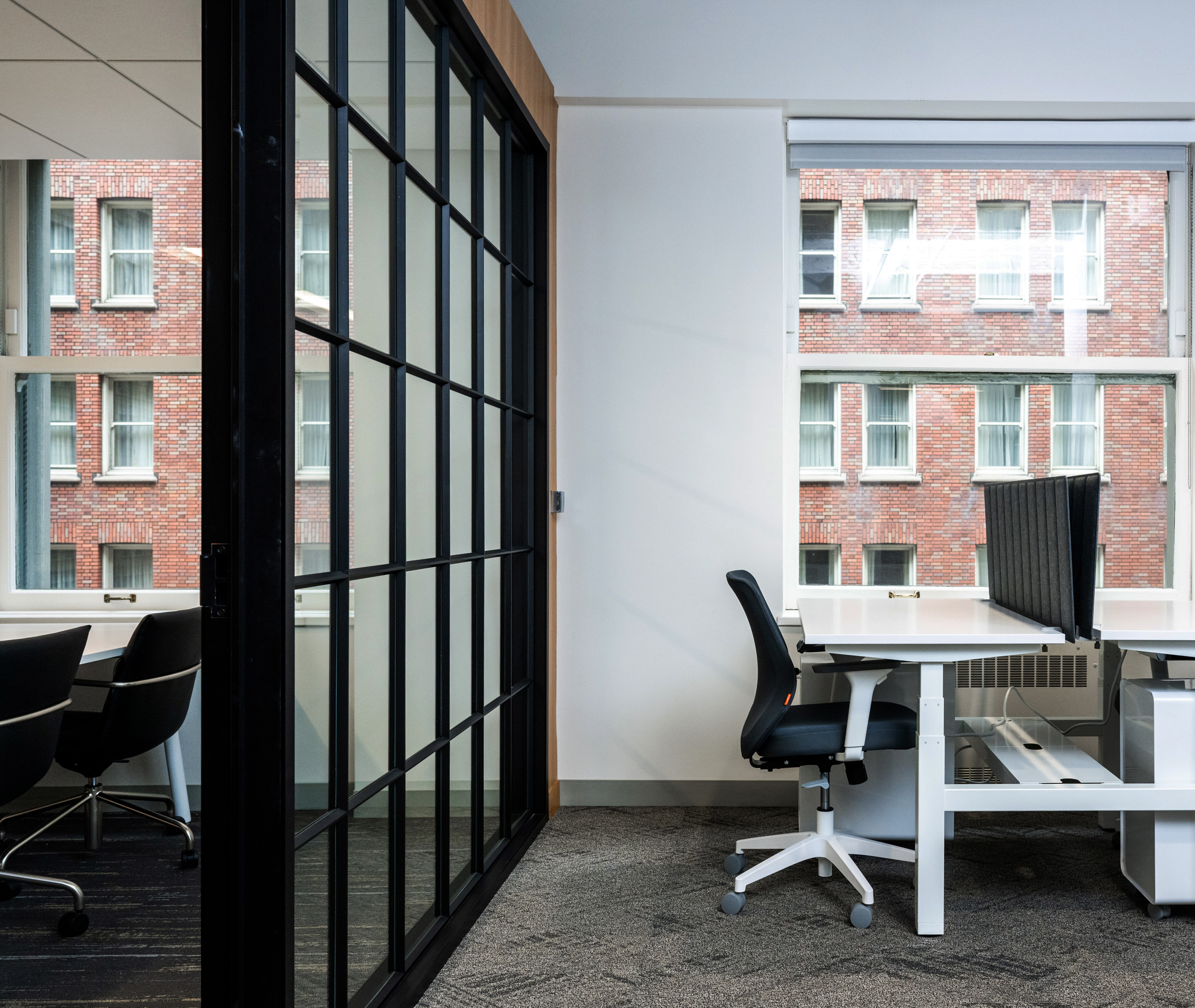 The image shows a modern office space with a black sliding glass partition. On the right is a desk with a black chair, and large windows overlook a brick building.