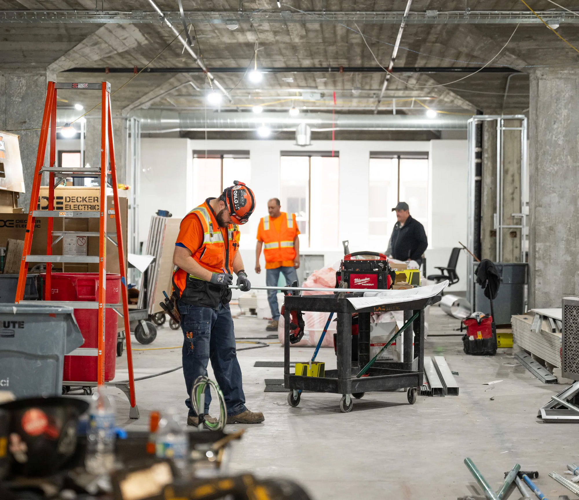 Workers in hard hats and vests are in a construction site, surrounded by tools and equipment. A man is focused on his task, with others in the background.