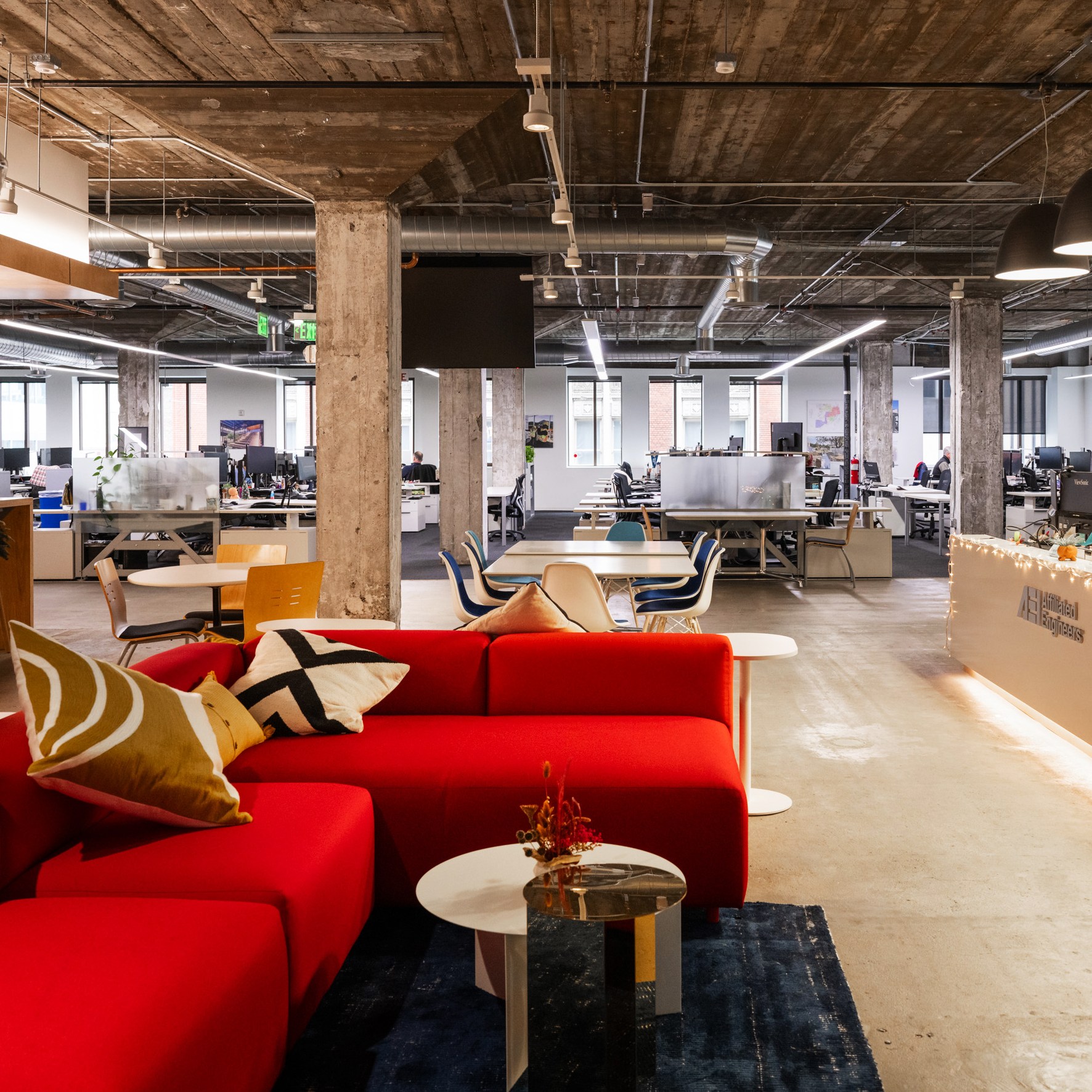 The image shows a modern office space with open seating, a red couch, a wooden reception desk, and industrial-style decor featuring exposed pipes and concrete beams.