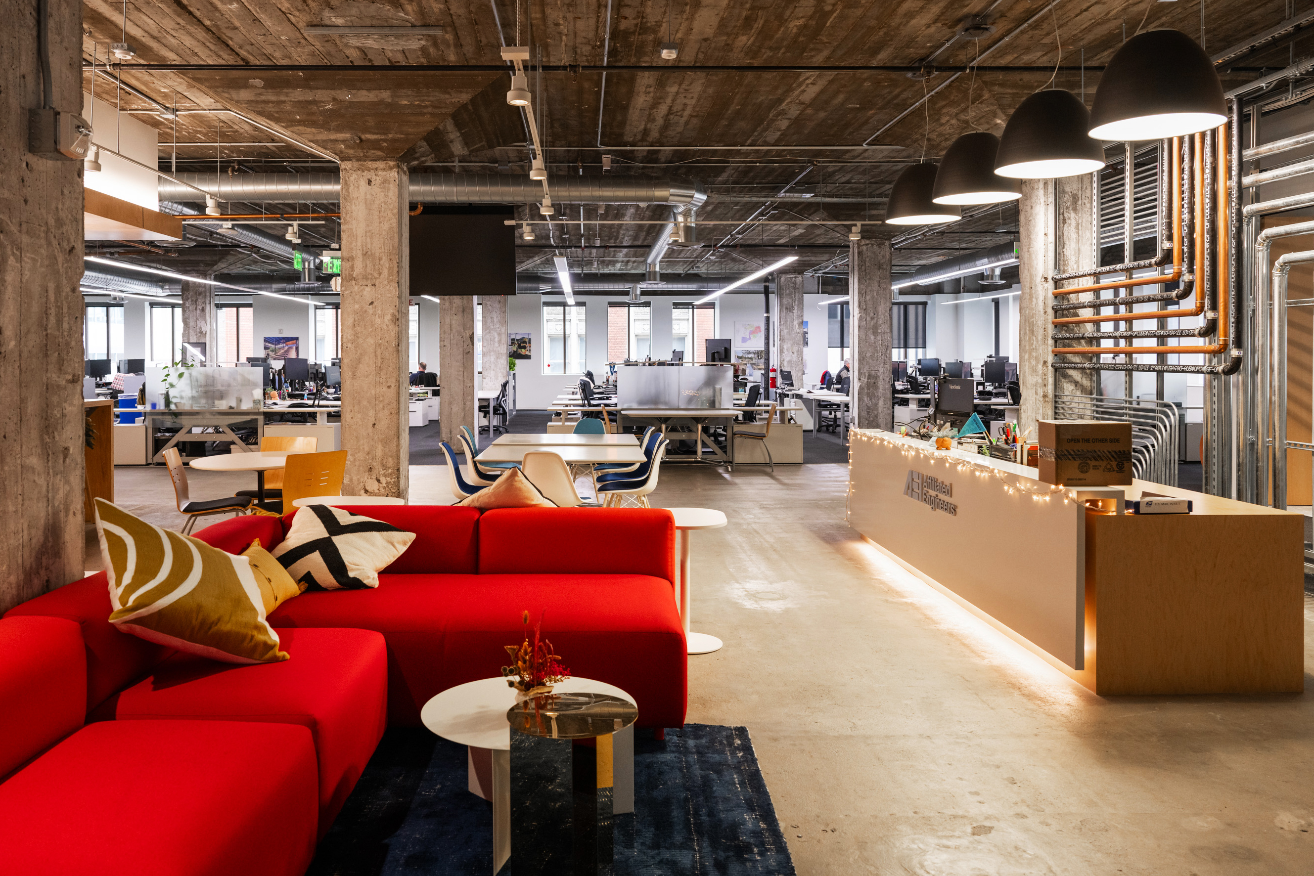 The image shows a modern office space with open seating, a red couch, a wooden reception desk, and industrial-style decor featuring exposed pipes and concrete beams.