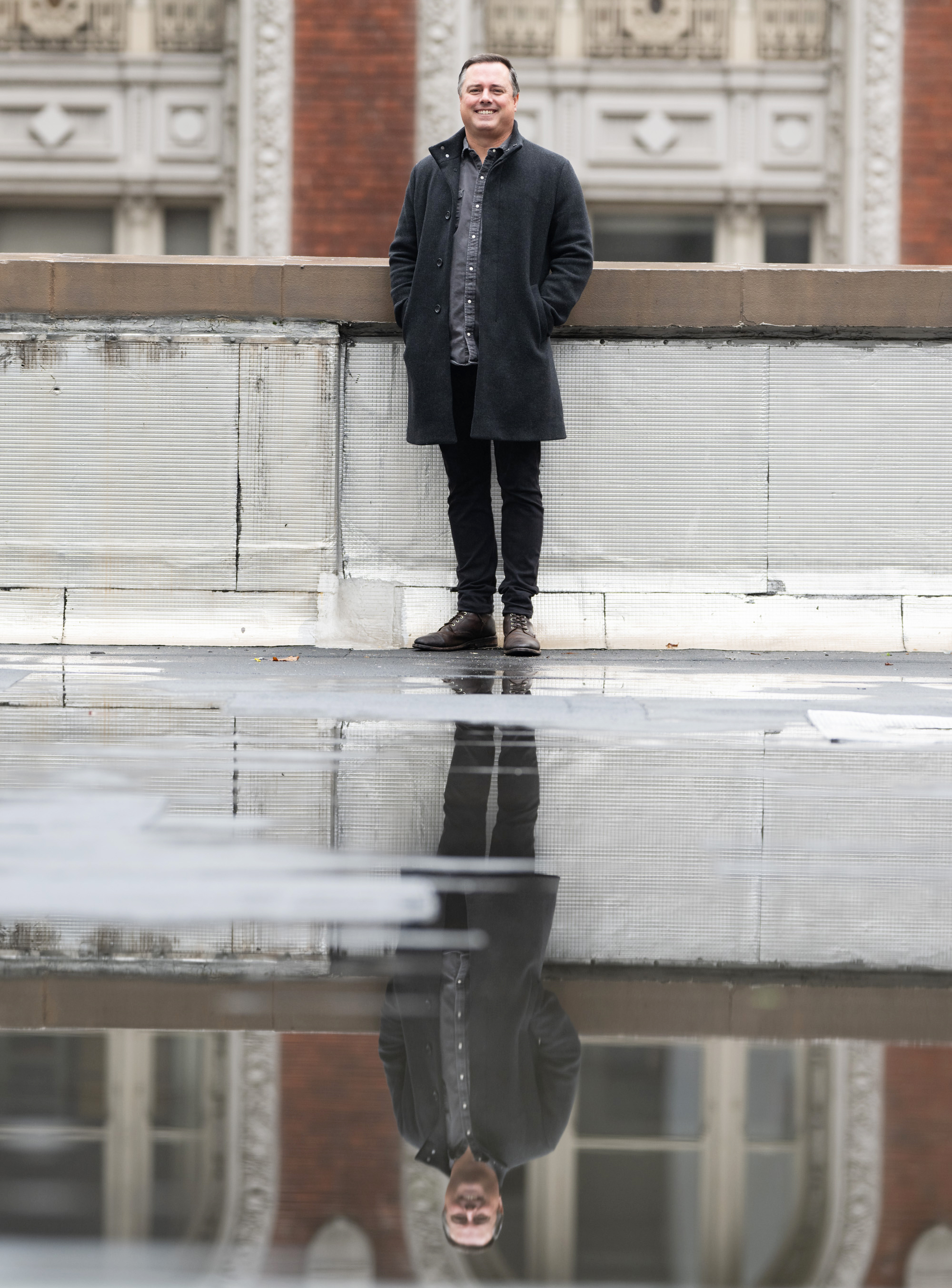 A man in a dark coat stands on a rooftop, with a blurred architectural background. His reflection is visible in a puddle on the ground.