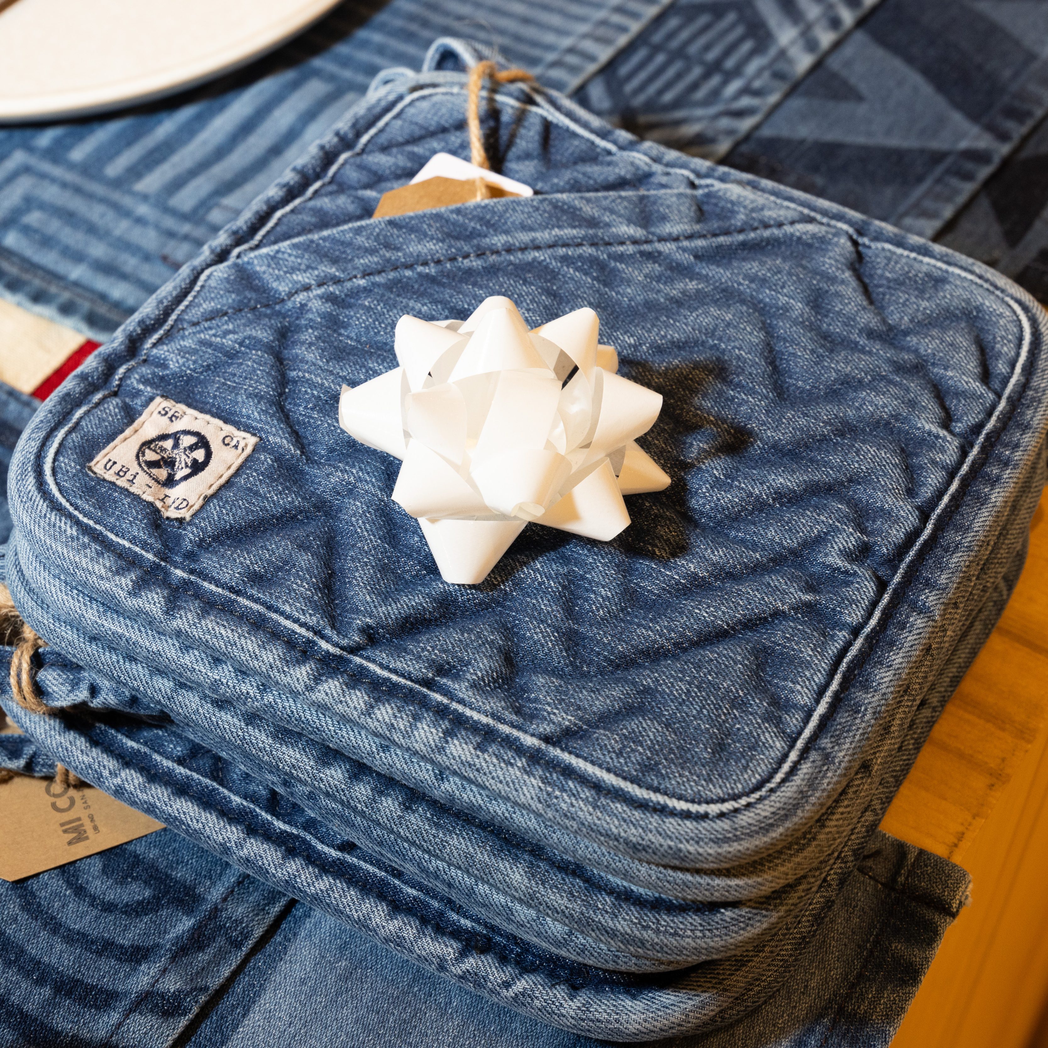 Denim pot holders with a patch and attached tags are stacked on a wooden surface. A white gift bow sits on top, next to a plate with cutlery.