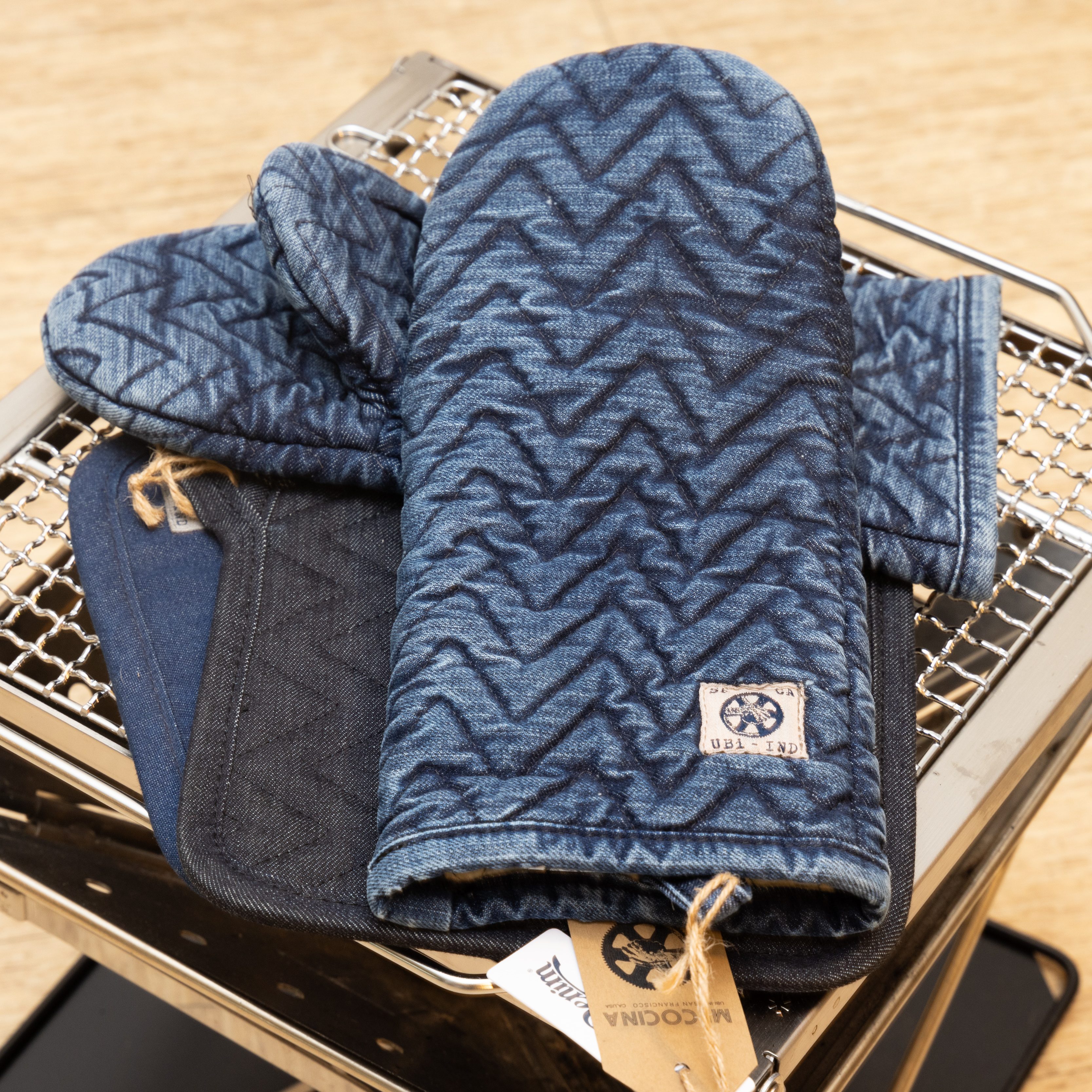 A blue quilted oven mitt with a zigzag pattern is placed on a metal rack. It has a label and twine loop attached to it.