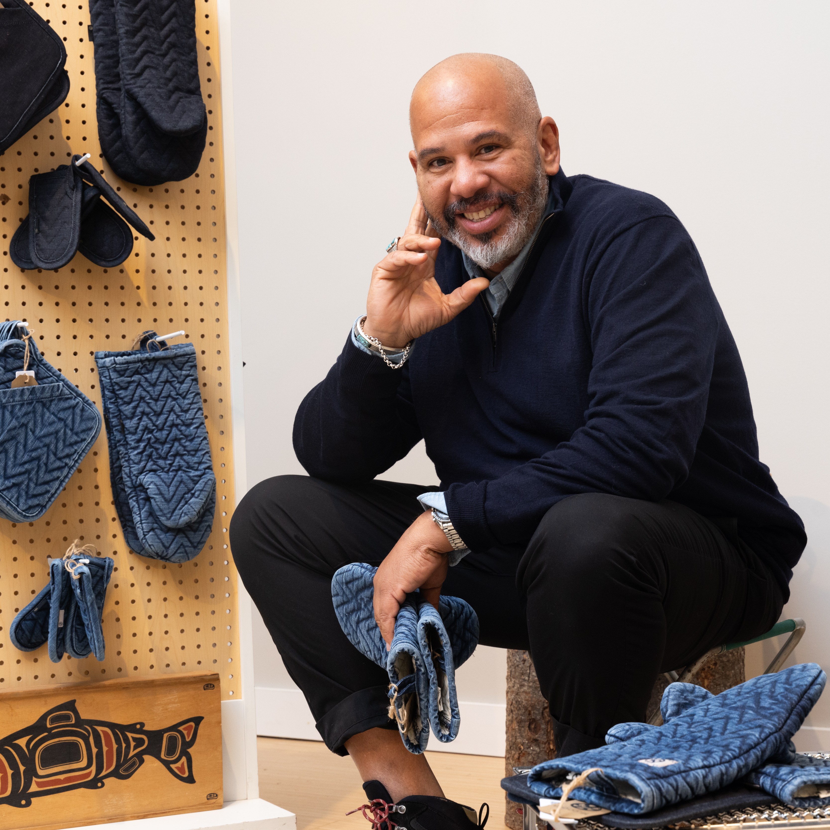 A man with a beard is smiling and crouching beside a pegboard displaying blue quilted kitchen items. There's also a wooden art piece of a fish.