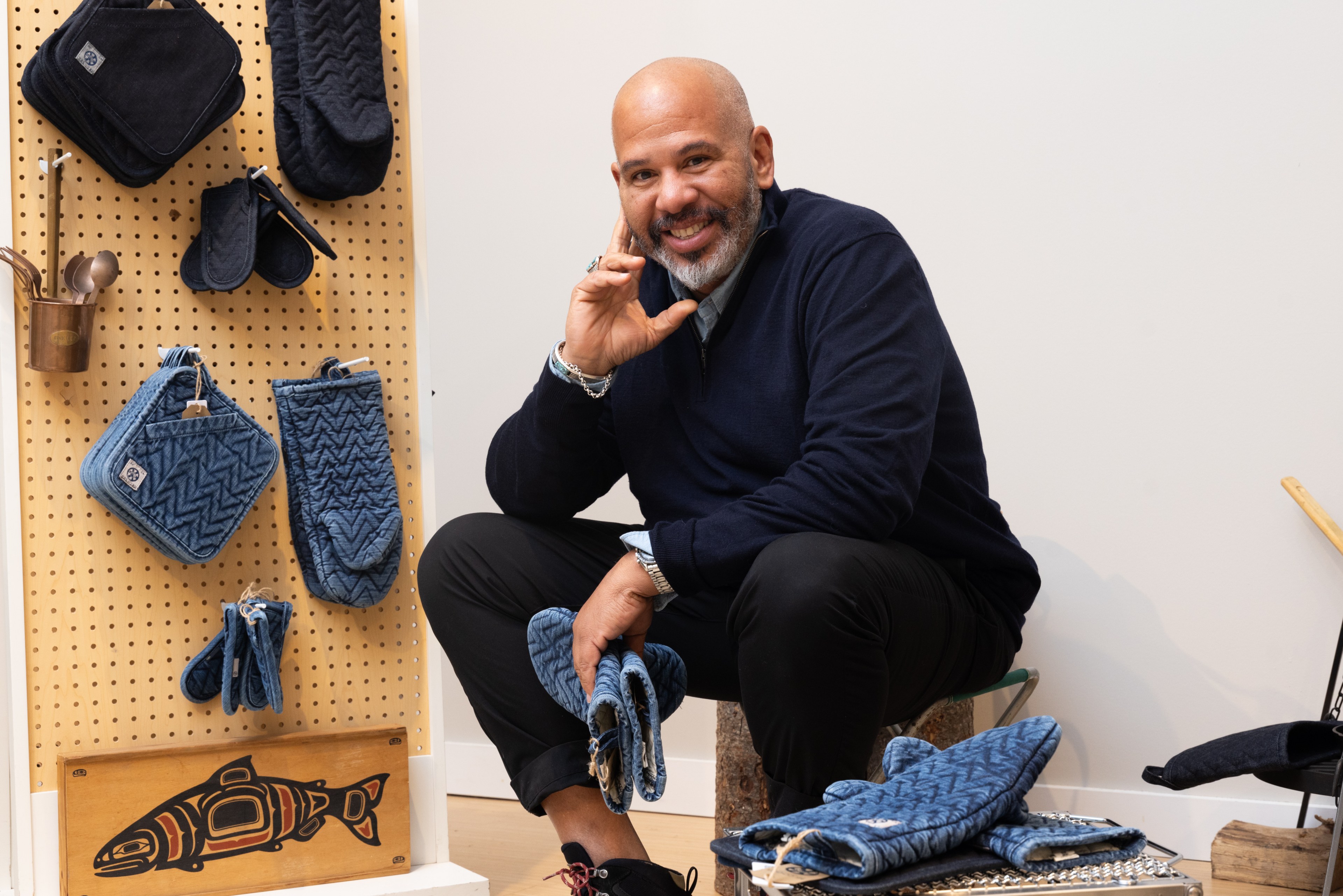 A man with a beard is smiling and crouching beside a pegboard displaying blue quilted kitchen items. There's also a wooden art piece of a fish.