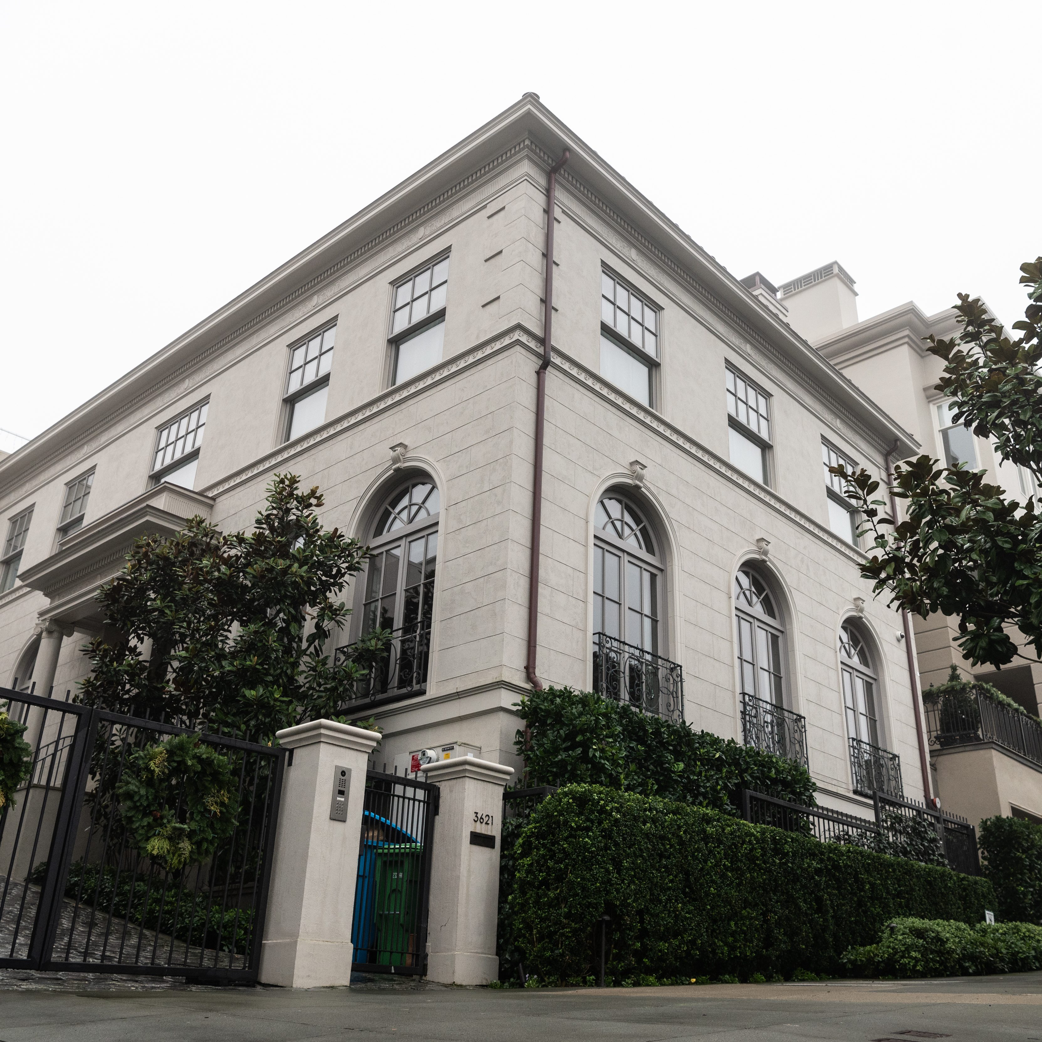 The image shows a large, elegant, cream-colored building with tall arched windows and black metal railings. It’s surrounded by greenery and enclosed by a black gate.