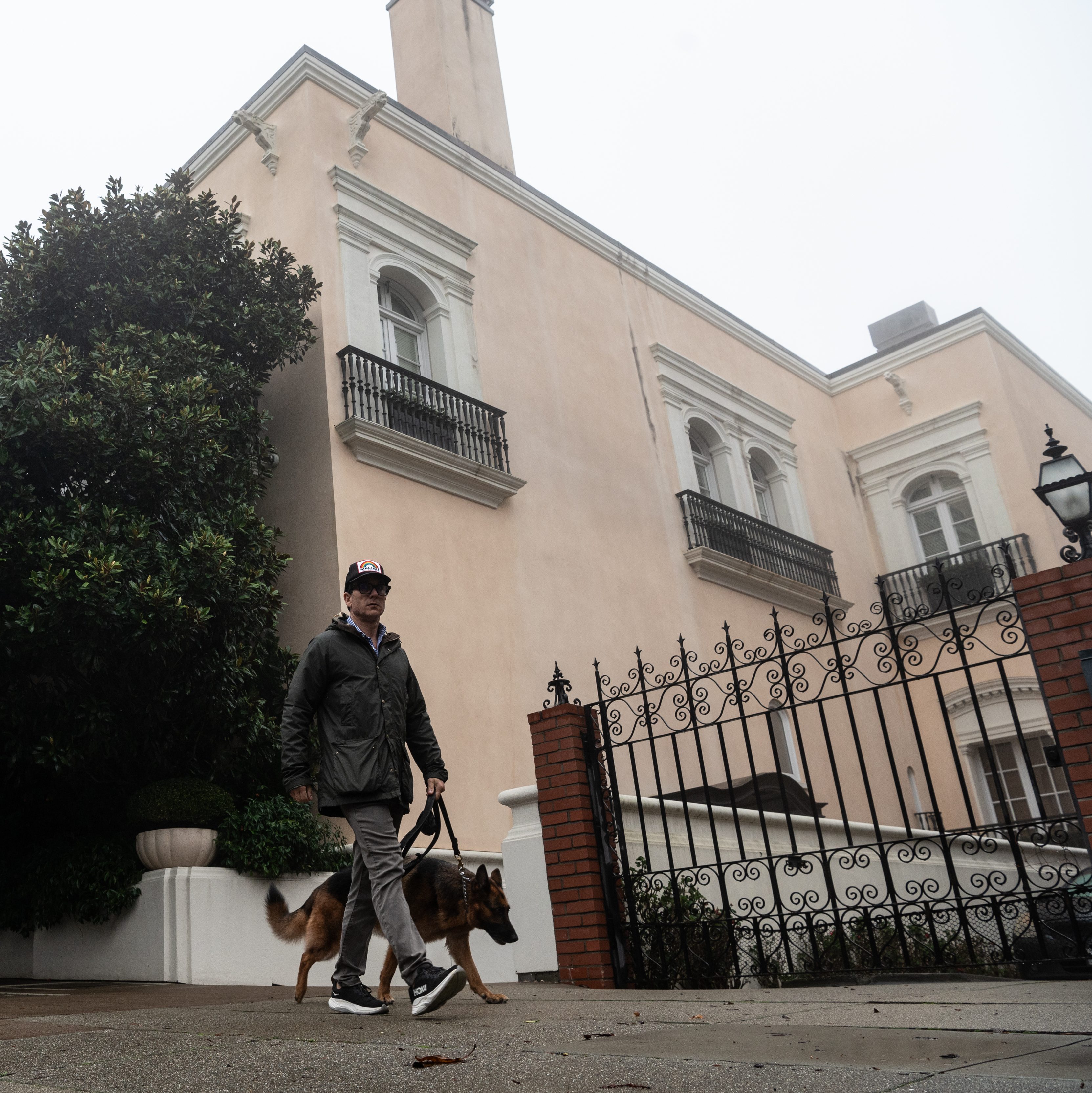 A man walks a German Shepherd past a large, light-colored house with black wrought-iron balconies and a gate, surrounded by greenery on a misty day.